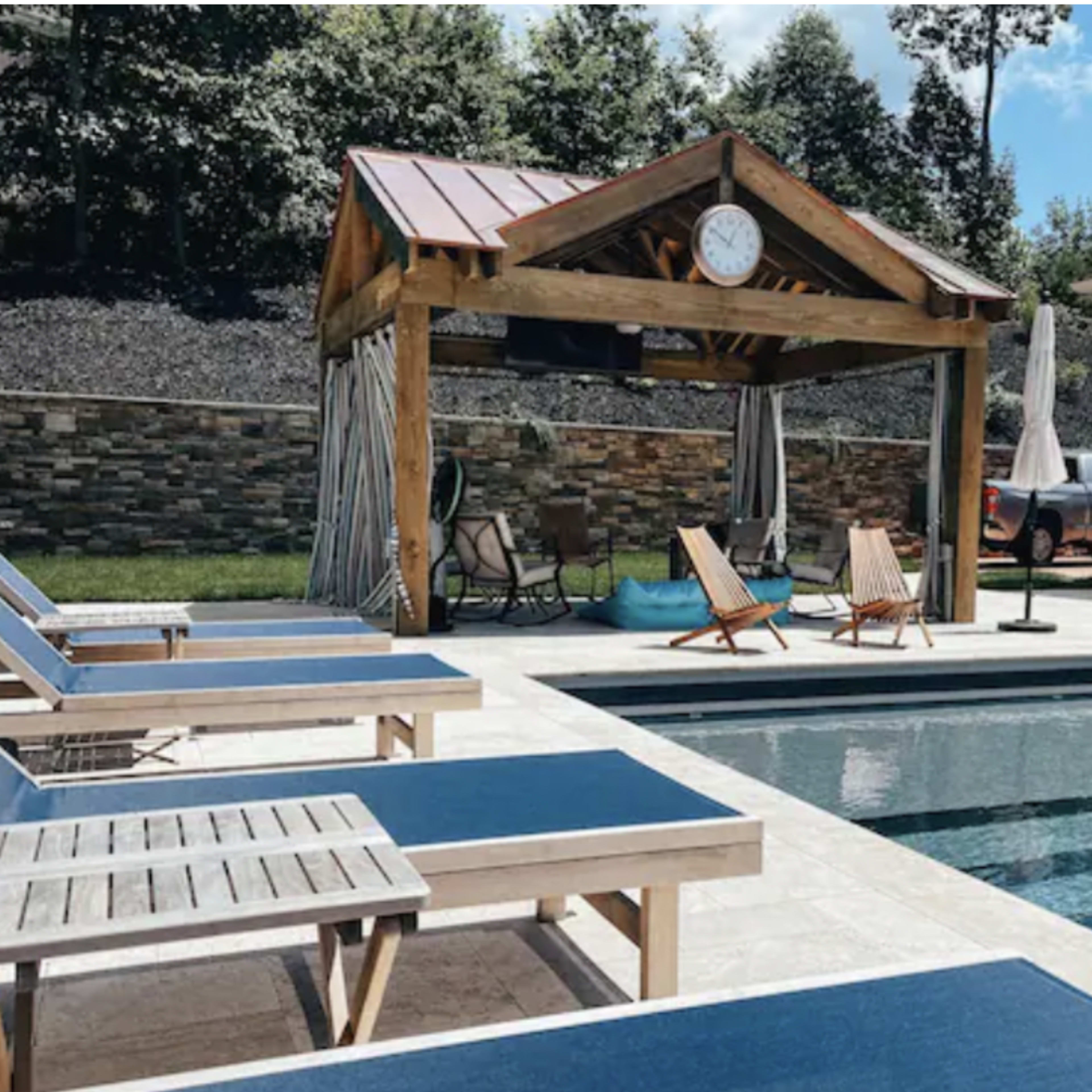 A swimming pool area features lounge chairs, a covered gazebo with a clock, and a stone wall backdrop.