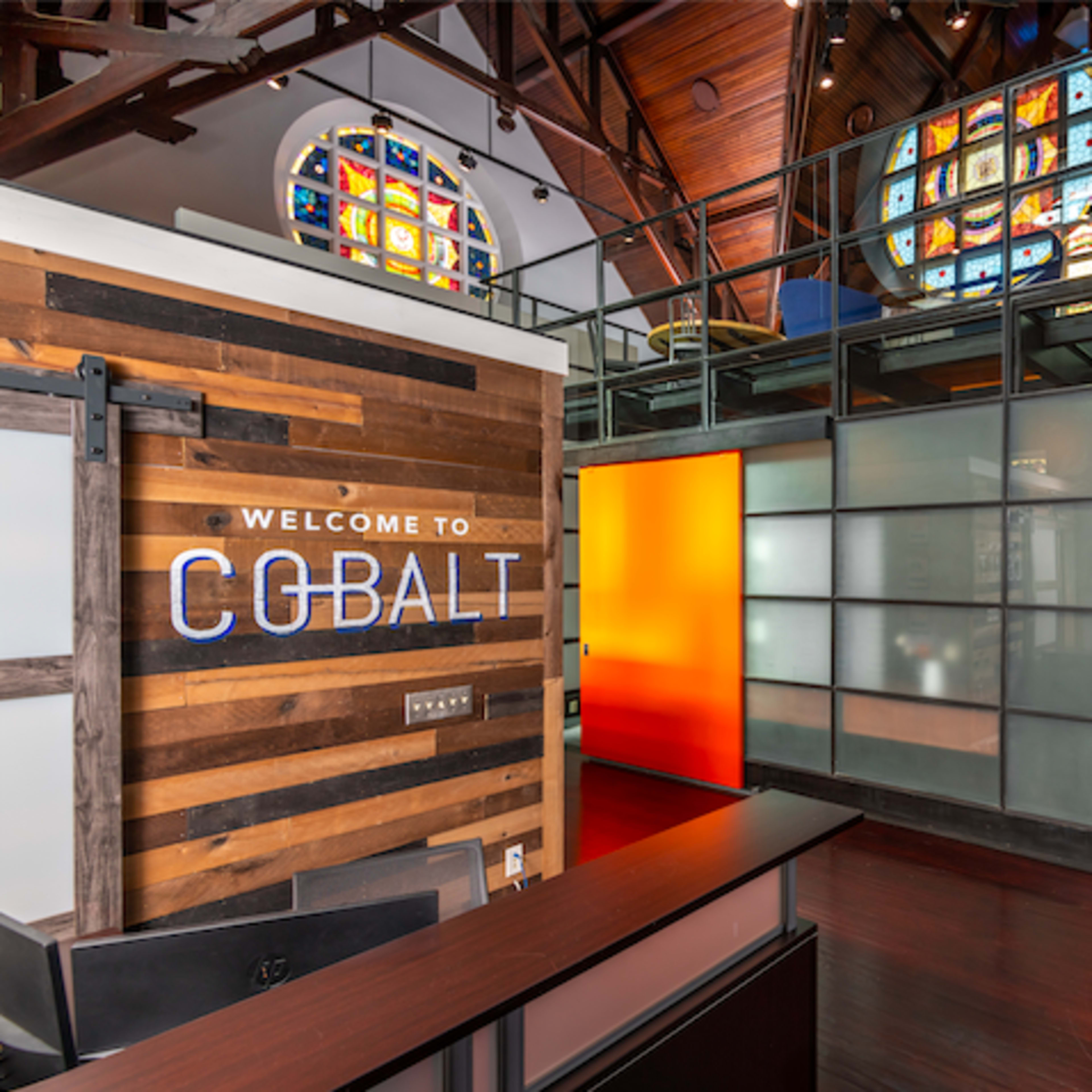 A modern office interior with a wooden reception area and glass-enclosed workspace, featuring colorful stained glass windows overhead.