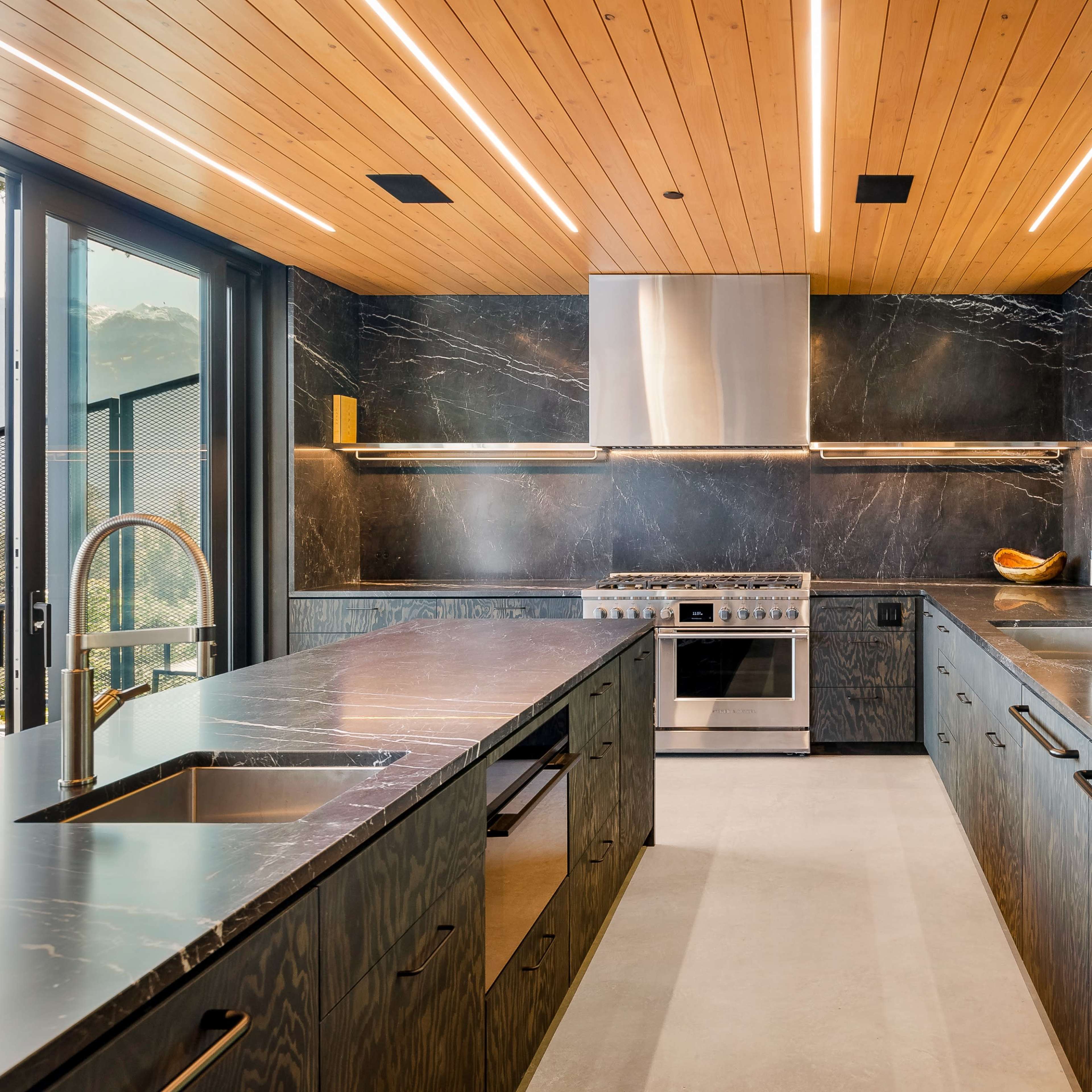 A modern kitchen with sleek black stone surfaces, wooden ceilings, and large windows overlooking greenery.
