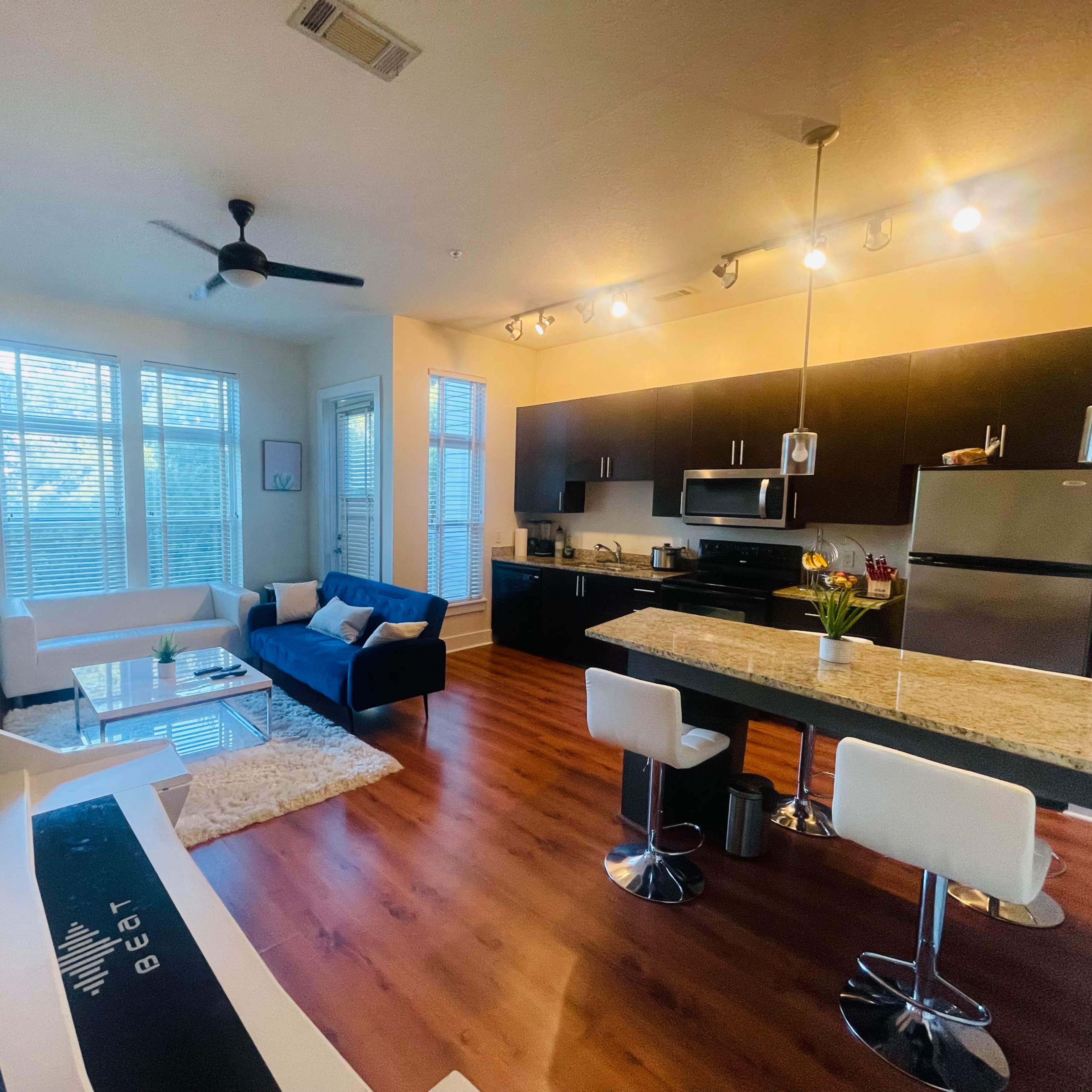 A modern apartment with a living area furnished with a white sofa, blue couch, and a kitchen featuring dark cabinets and a granite countertop.