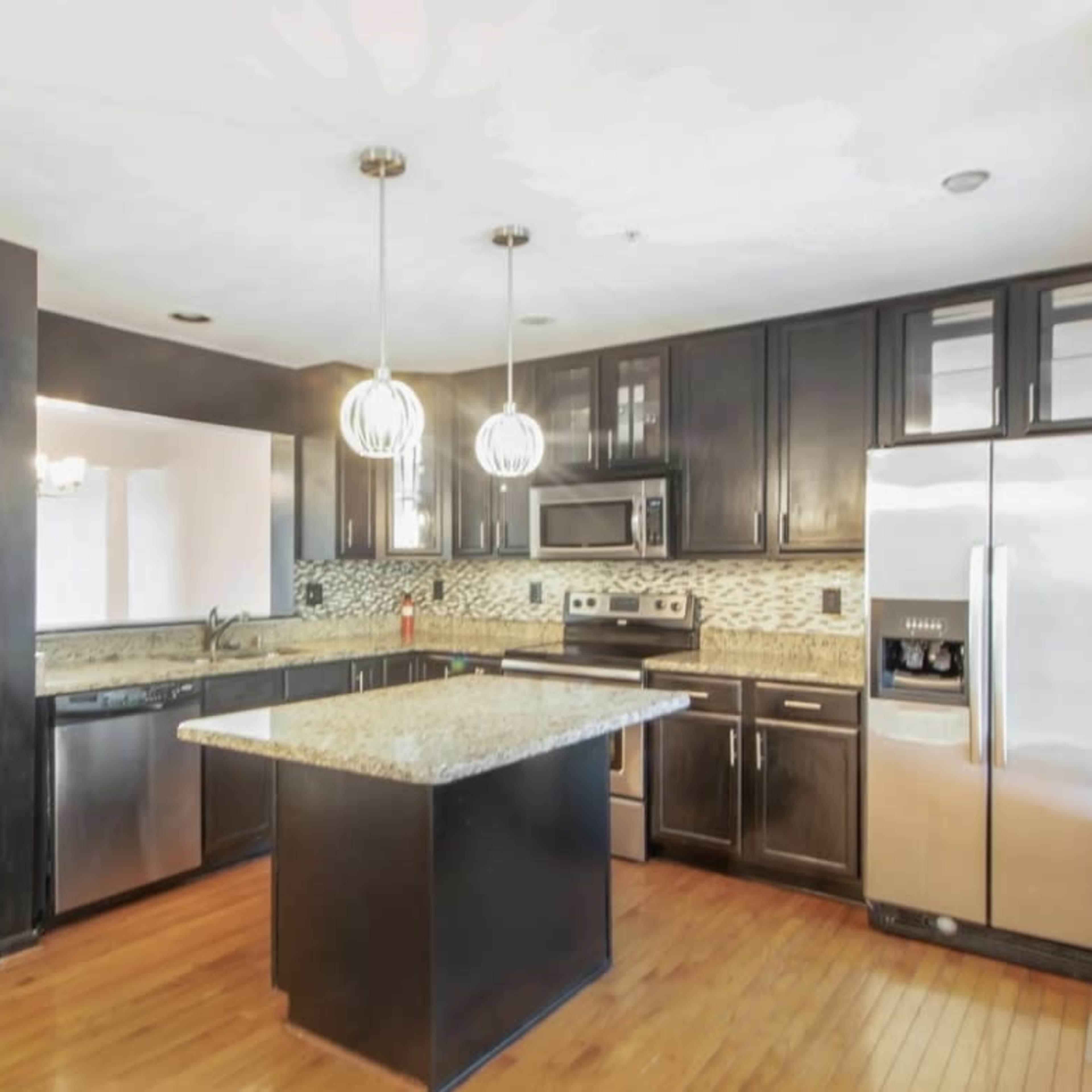 A modern kitchen with dark cabinetry, stainless steel appliances, and an island under pendant lighting.