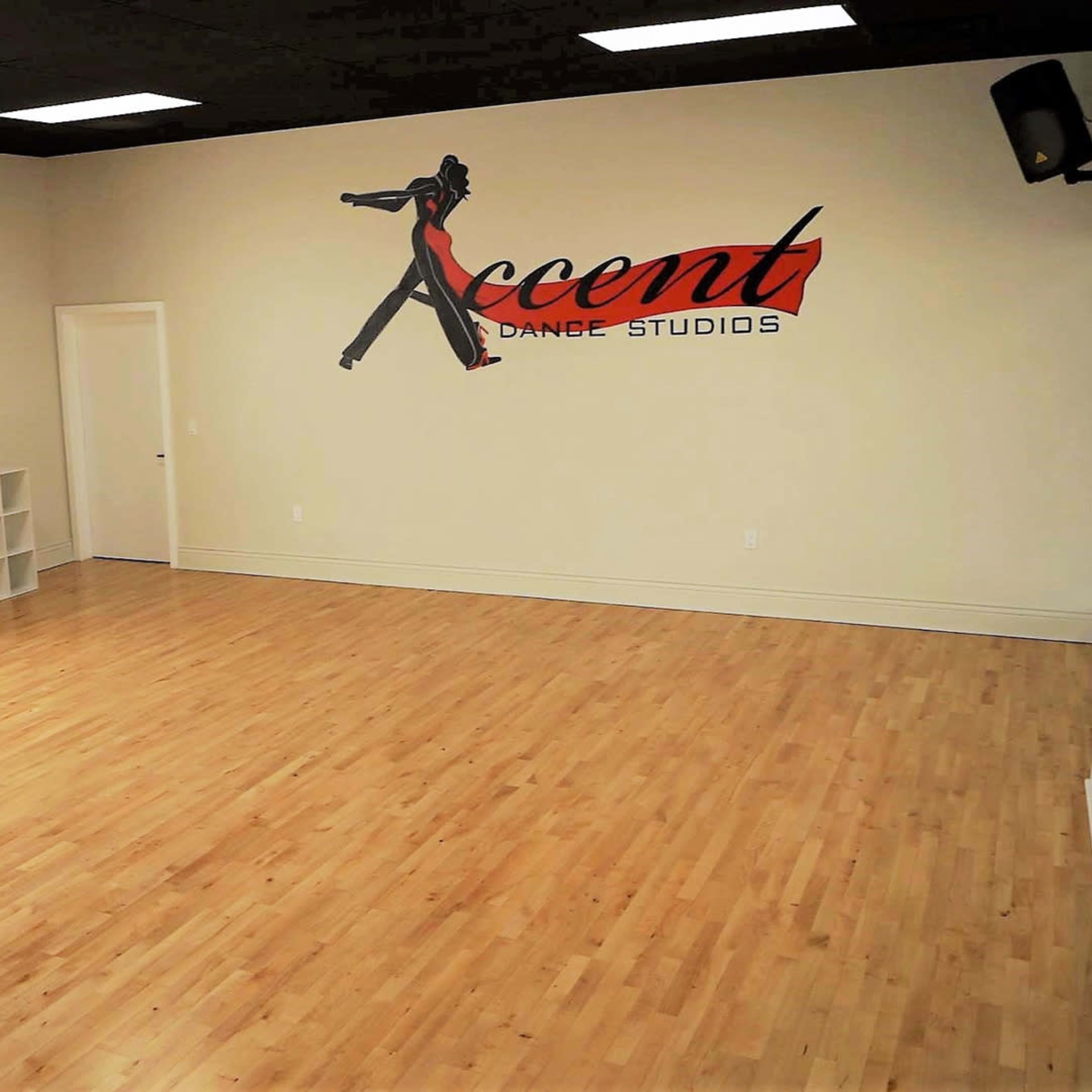 An empty dance studio with wooden floors, a large window, and a wall featuring a sign that reads "Accent Dance Studios."