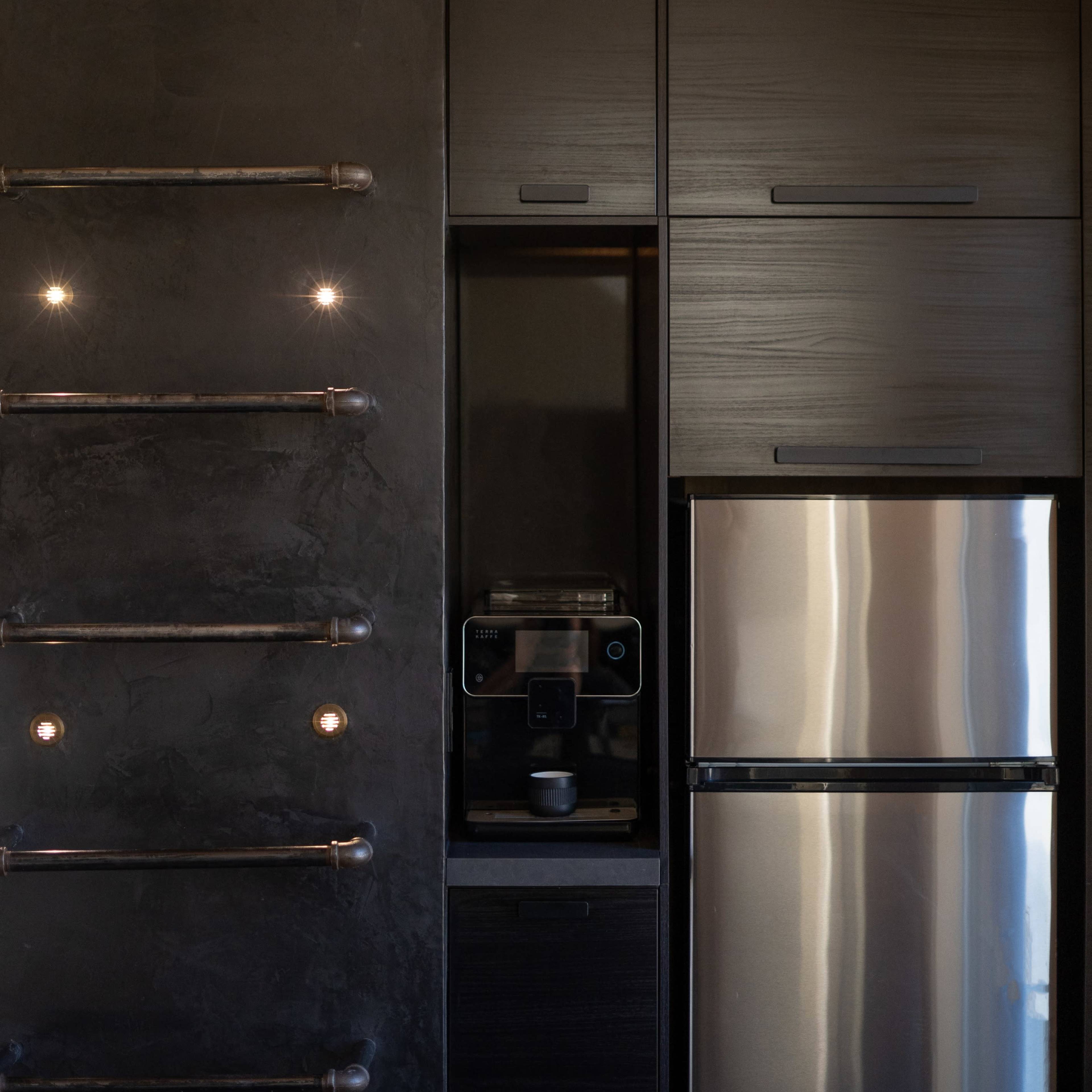 A modern kitchen features a stainless steel refrigerator and a built-in coffee maker against a dark wall with industrial-style light fixtures.