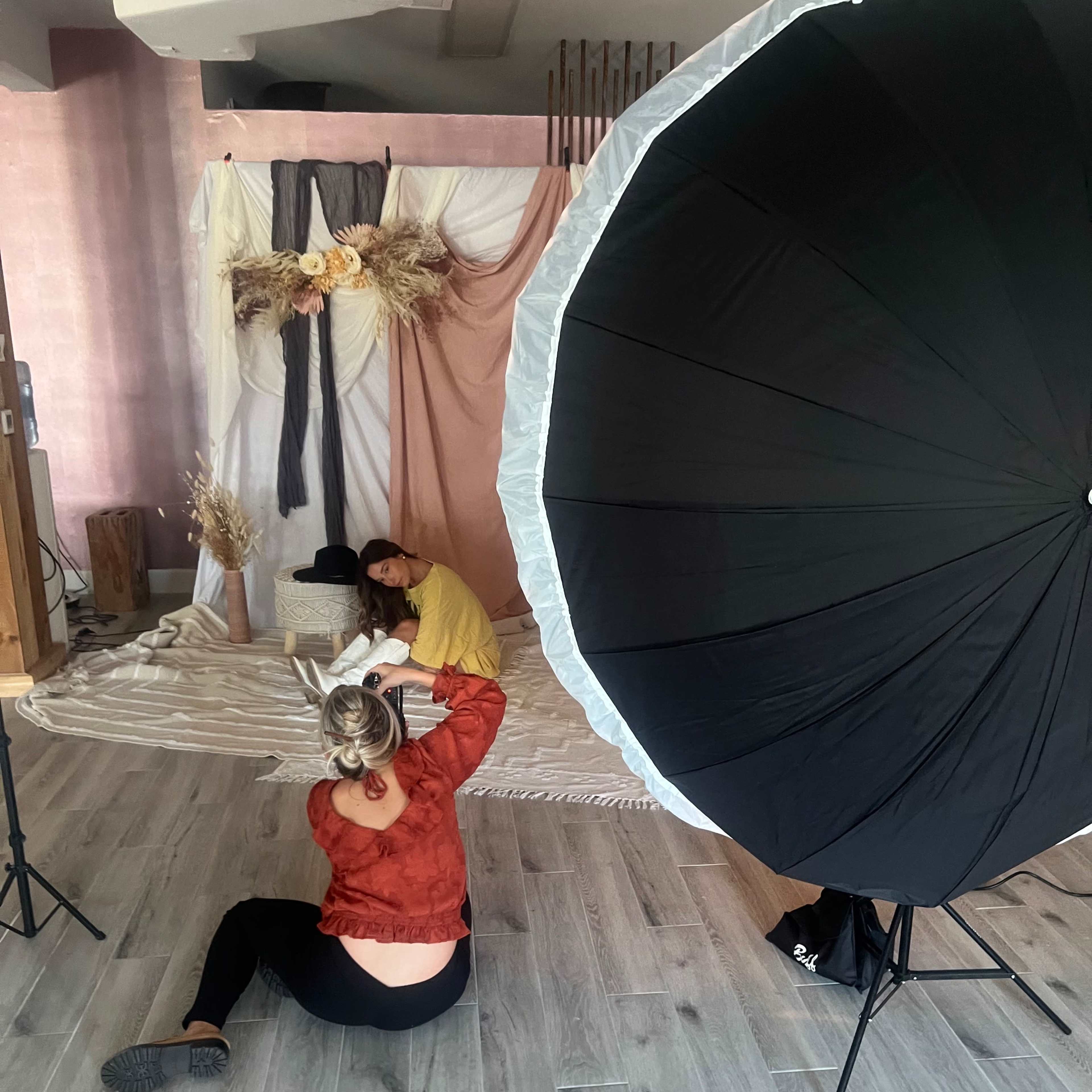 A photographer adjusts a model's pose in front of a decorated backdrop while using a large softbox for lighting.