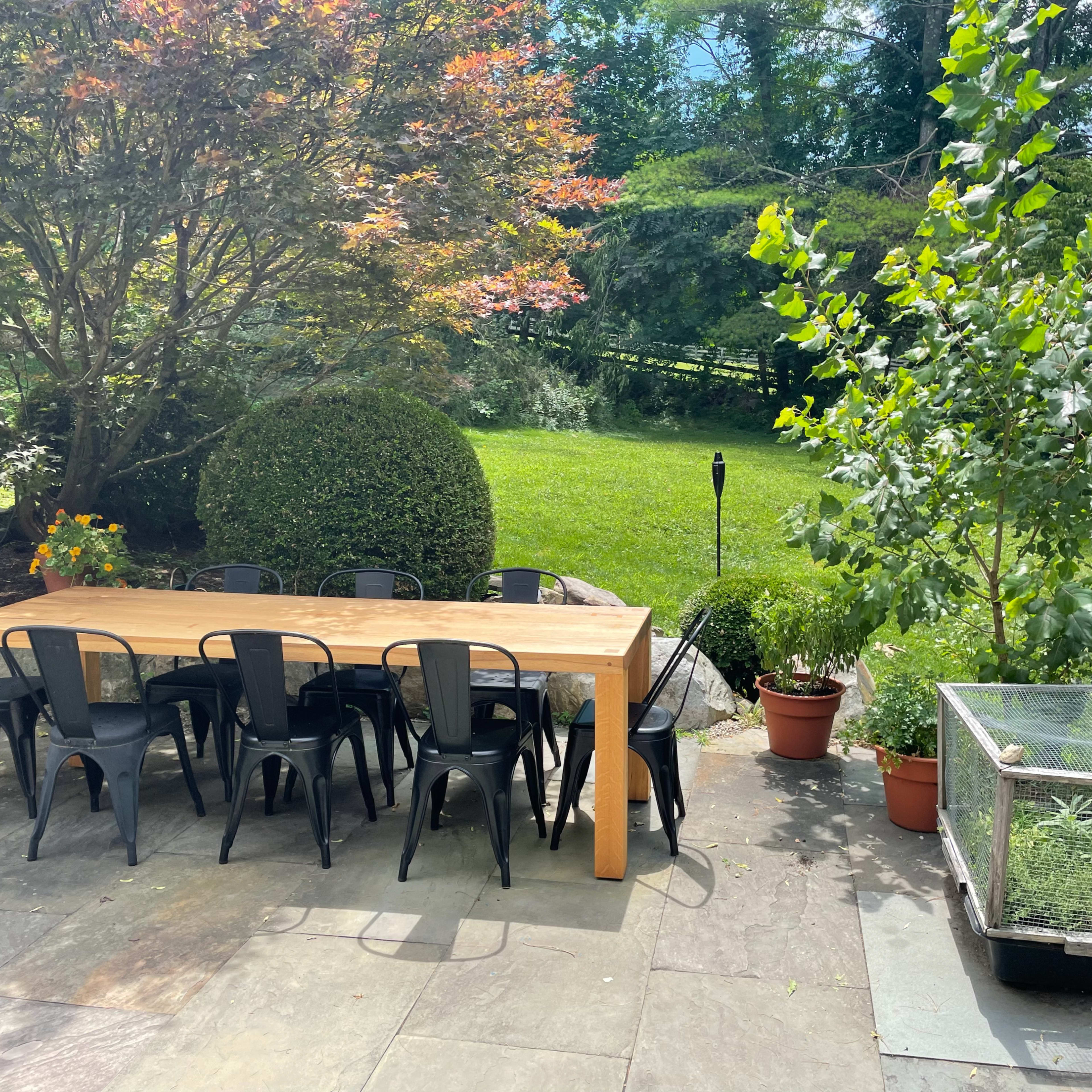 A long wooden dining table with black chairs is set on a stone patio, surrounded by greenery and potted plants.