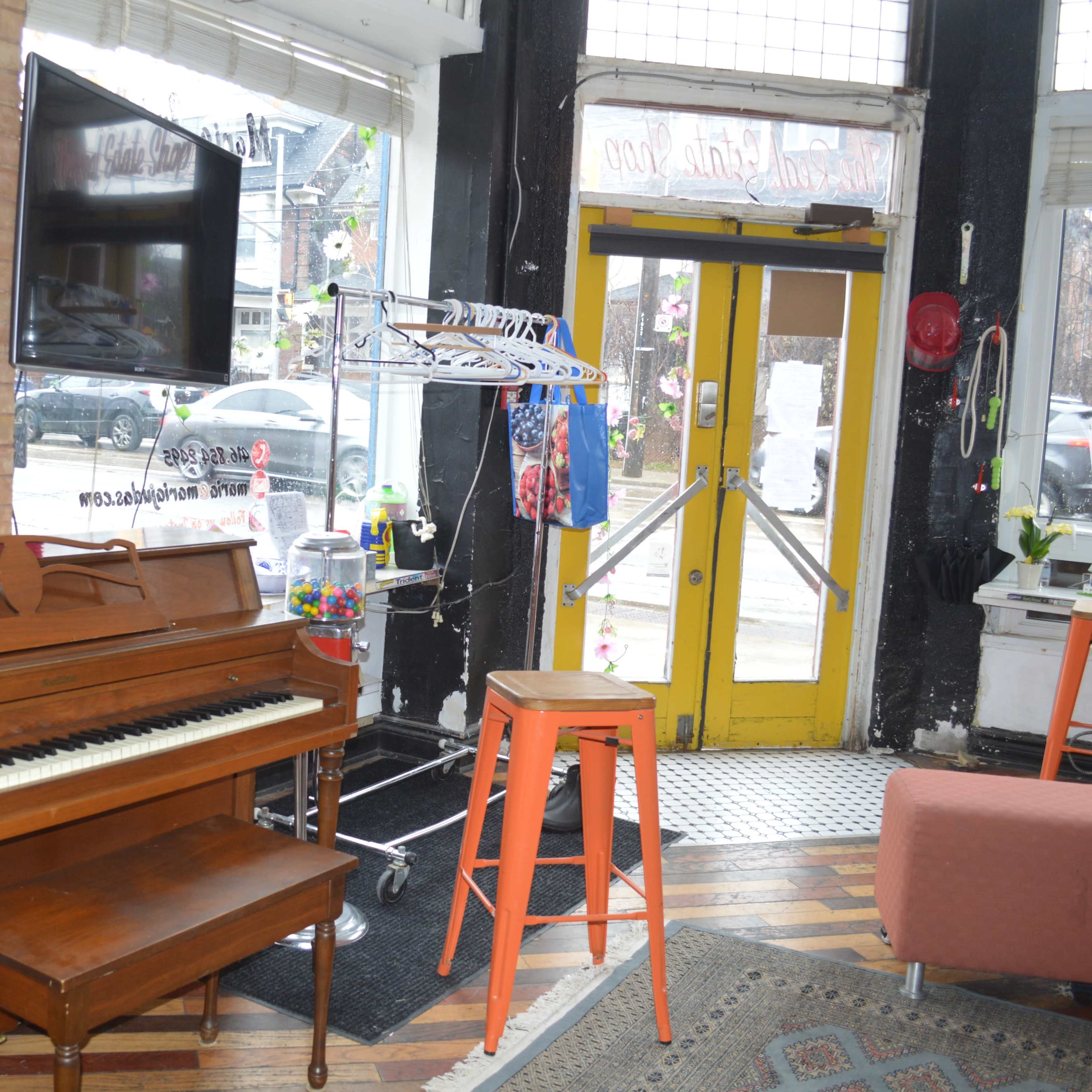 A café interior featuring a wooden piano, a television on the wall, and a yellow door leading outside, with seating and a clothing rack in the background.