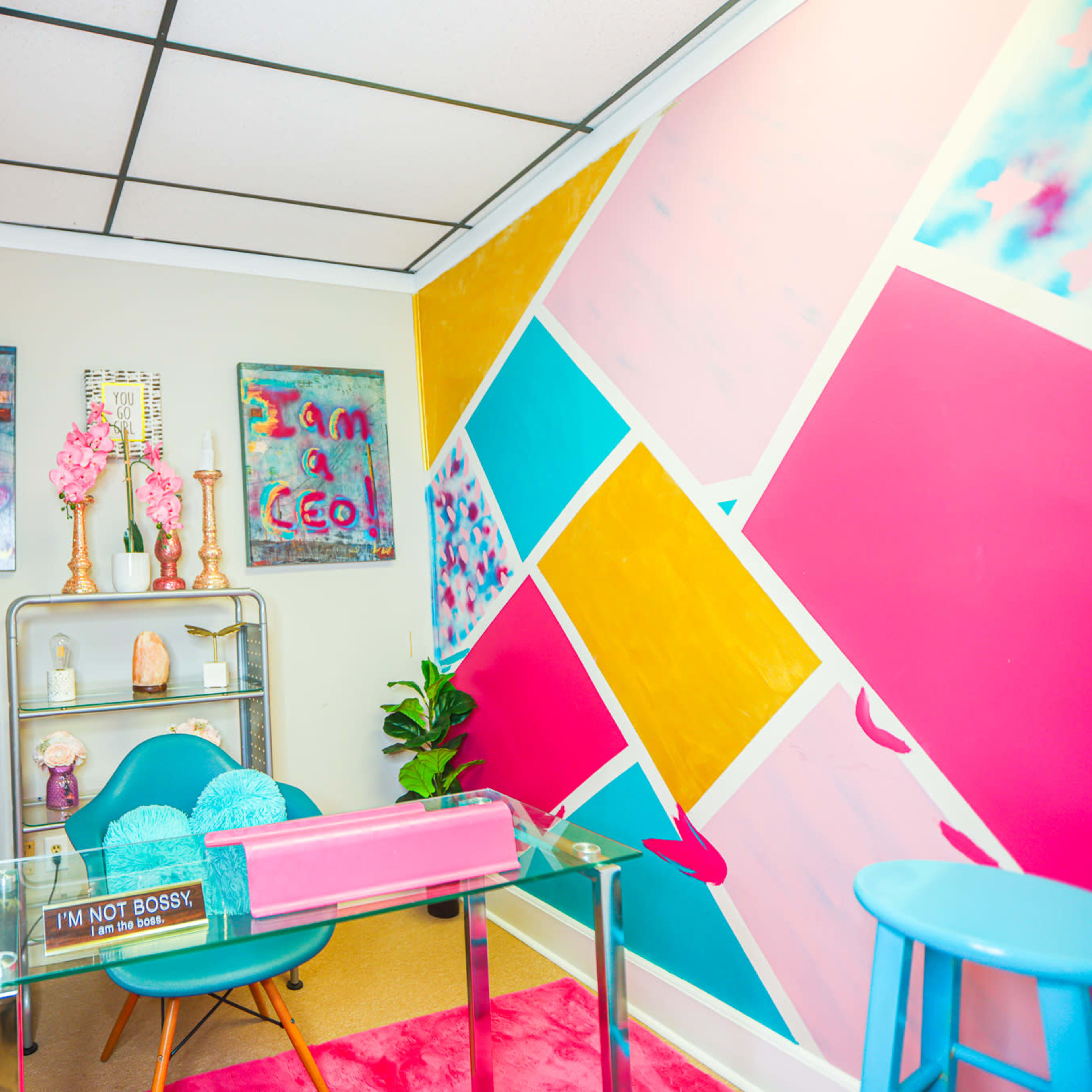 A small office space featuring a brightly colored accent wall, a glass desk with a pink and clear design, and decorative elements like artworks and plants.