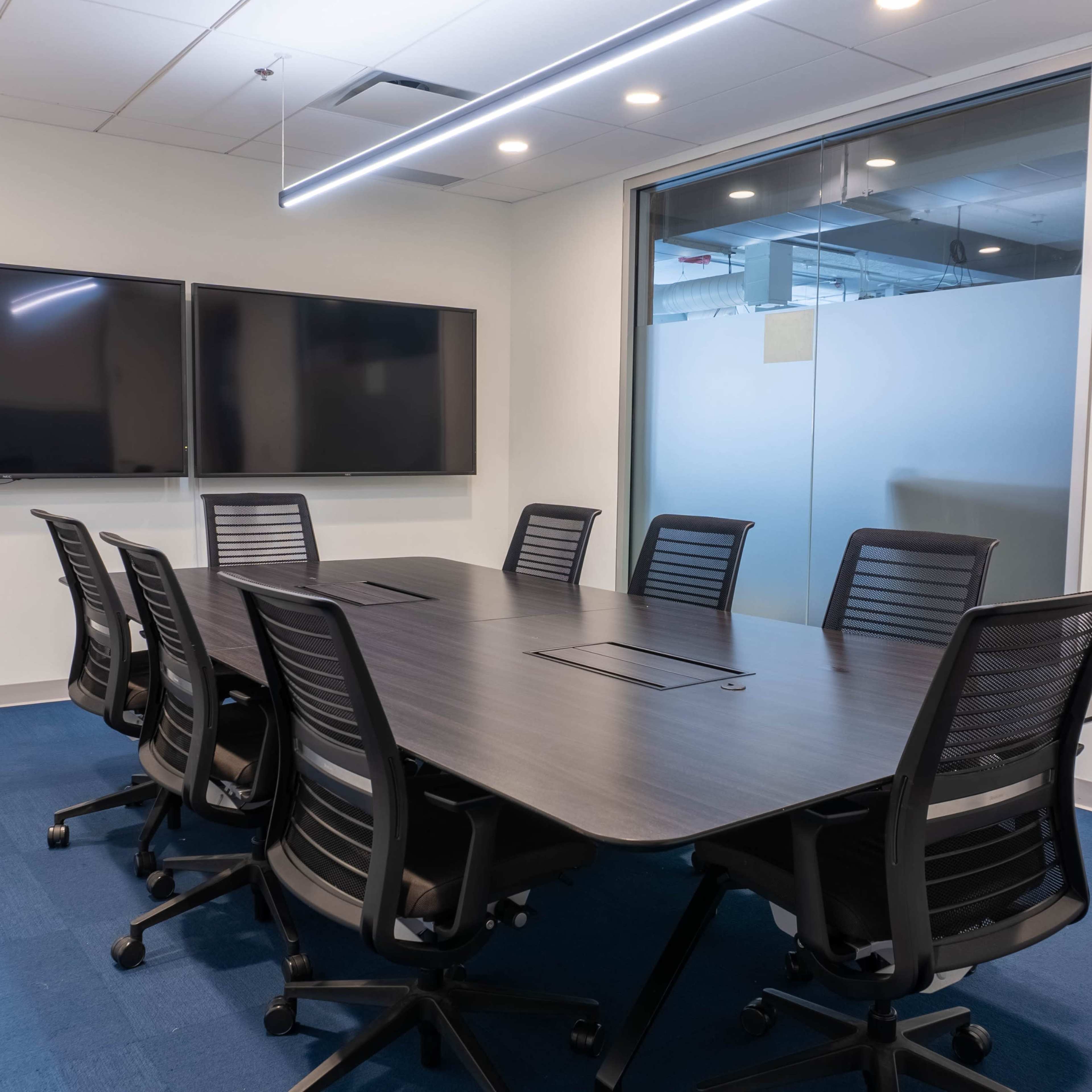 A modern conference room with a large dark table, eight rolling chairs, and two wall-mounted screens.