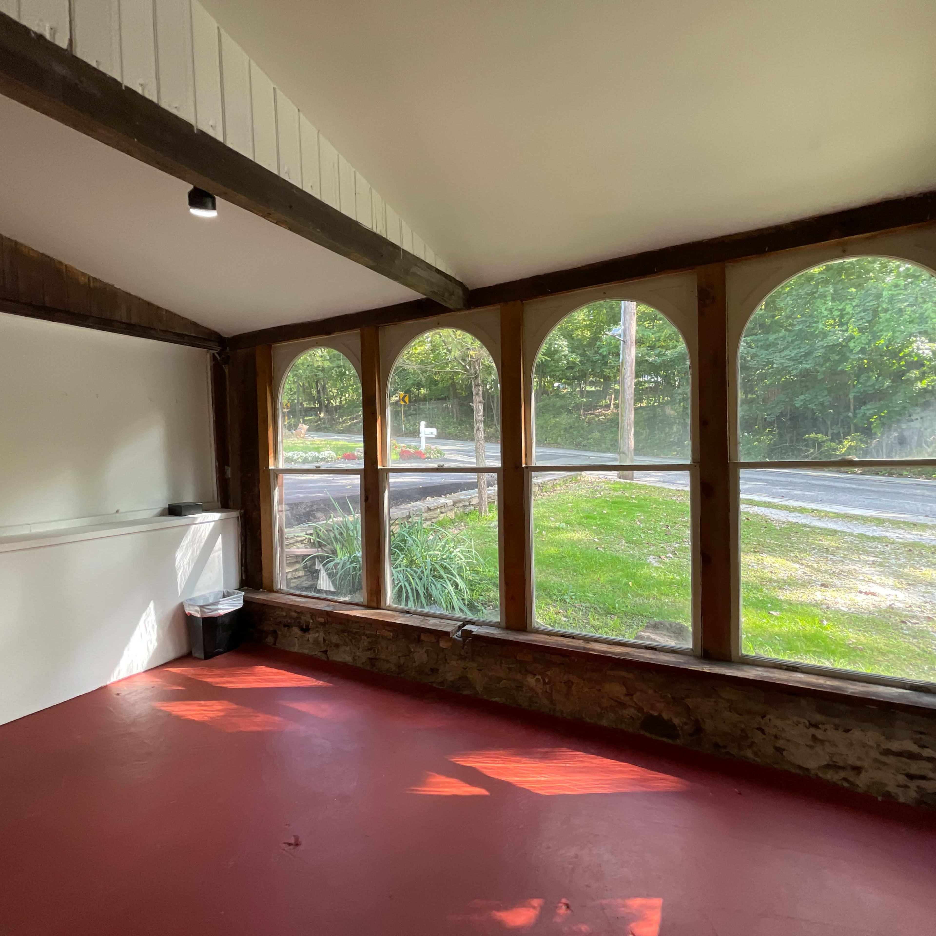 An empty, sunlit room with large arched windows overlooking a grassy area and a road outside.