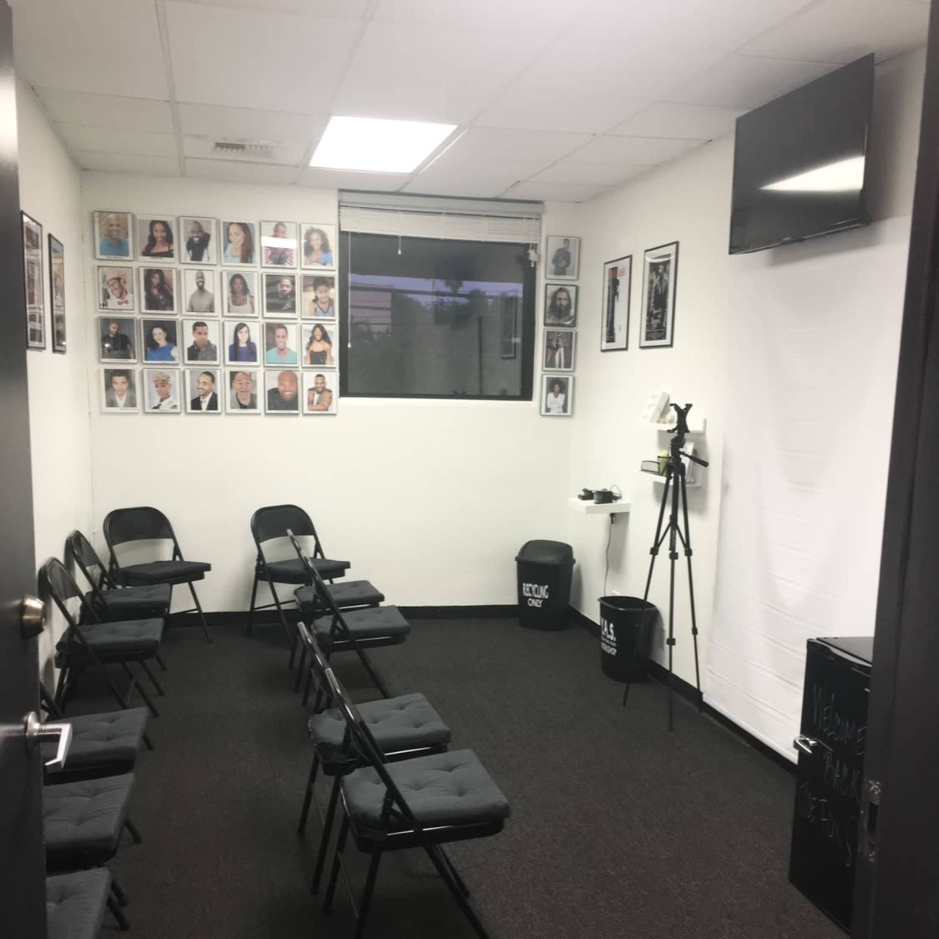 A small room with rows of black folding chairs facing a blank wall and a television, adorned with framed photographs on the adjacent wall.