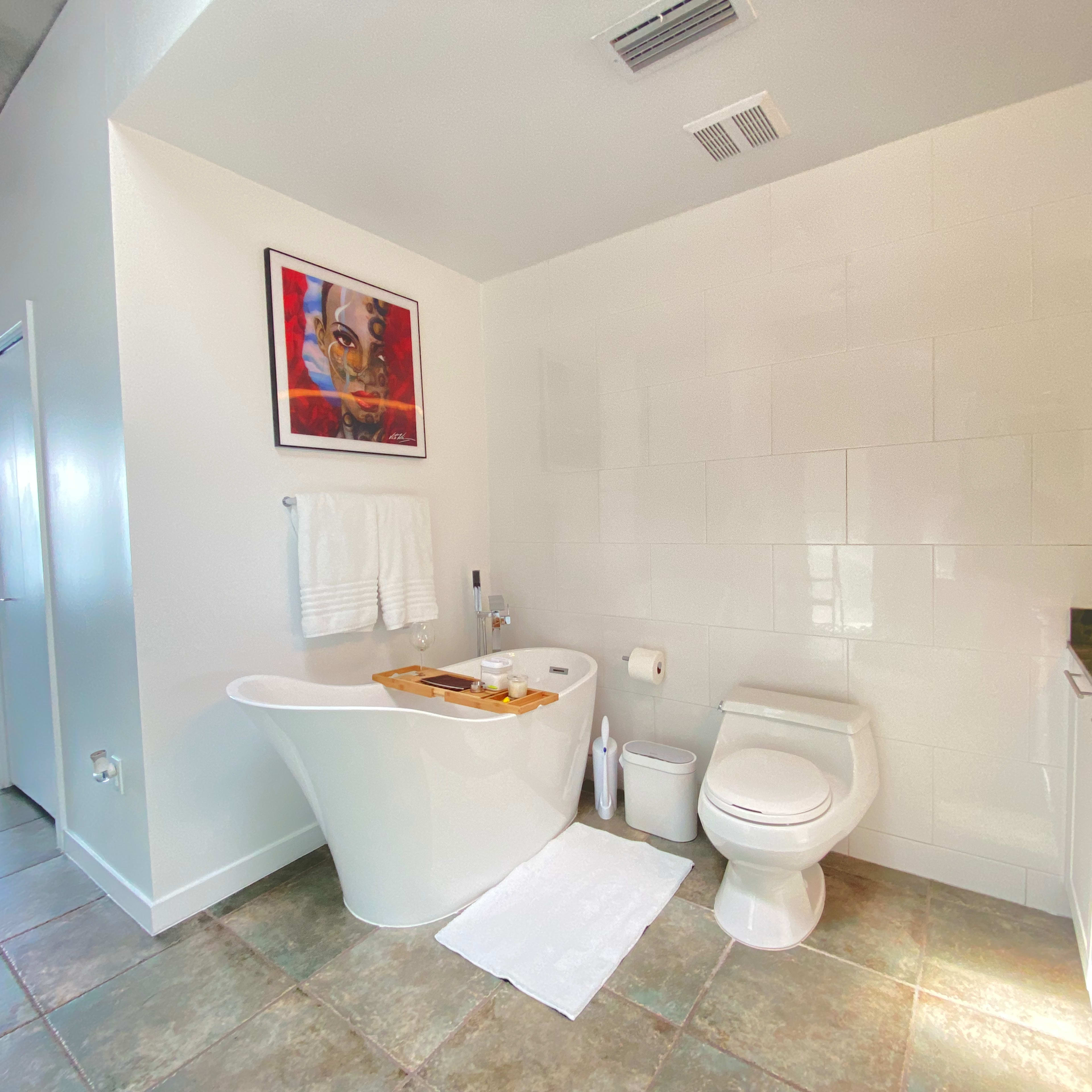 The bathroom features a freestanding bathtub with a wooden tray, a toilet, and a sink with a large mirror, all set against white tiled walls and a tiled floor.