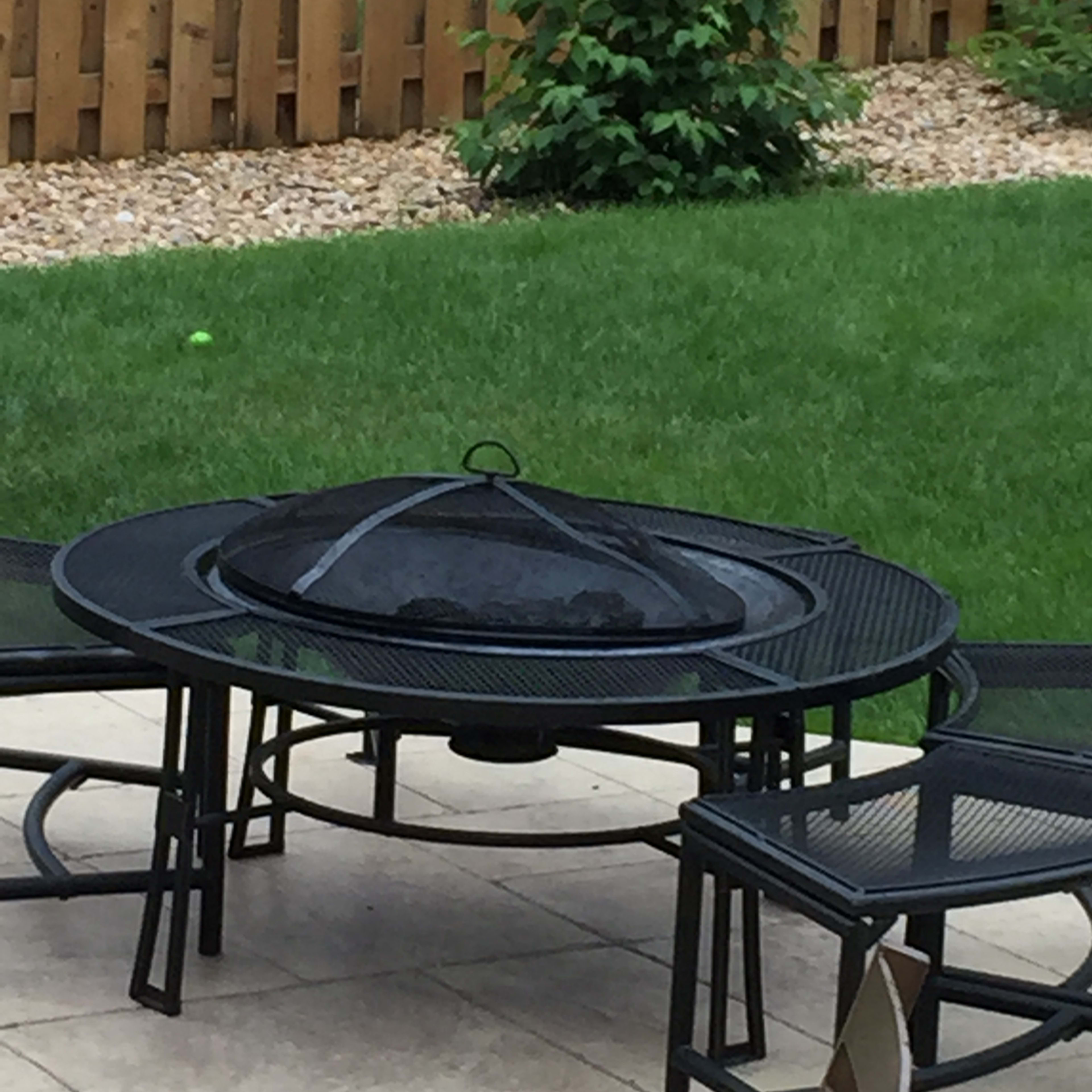 A black metal fire pit surrounded by chairs on a stone patio, with grass and a wooden fence in the background.
