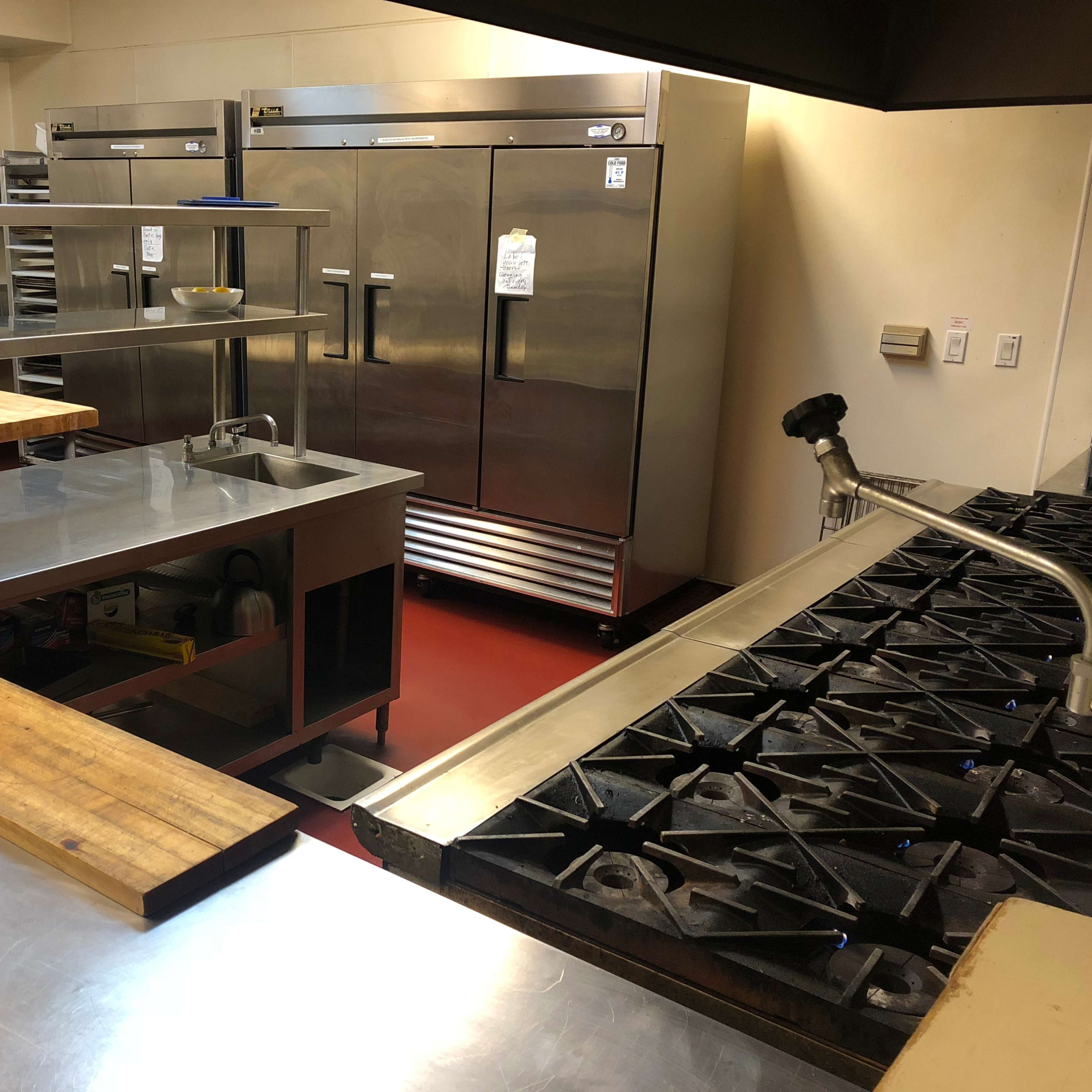 A commercial kitchen with stainless steel countertops, a large gas stove, and multiple refrigerators in the background.