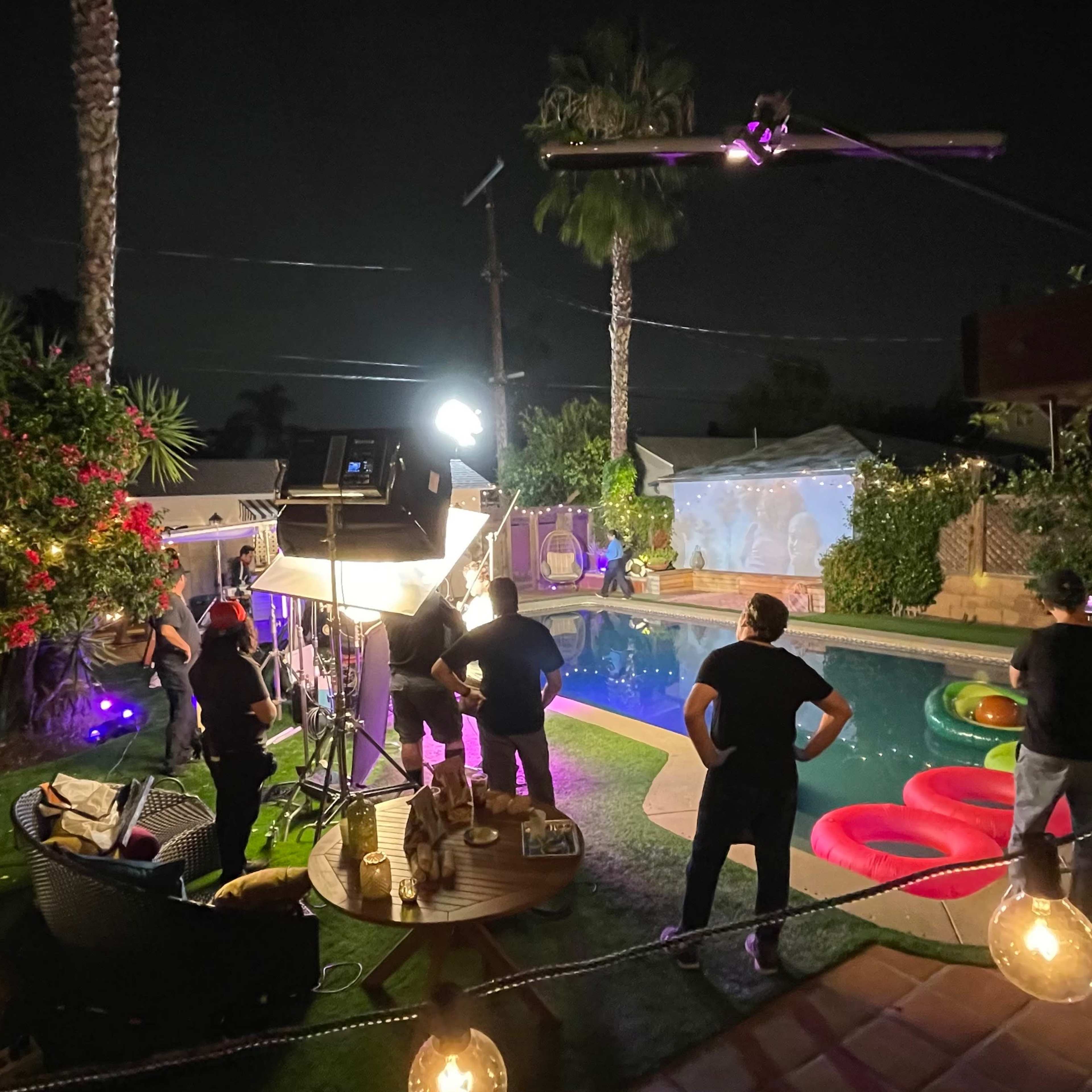 A film crew sets up lights and equipment around a swimming pool in a backyard decorated with palm trees and colorful inflatable floats at night.