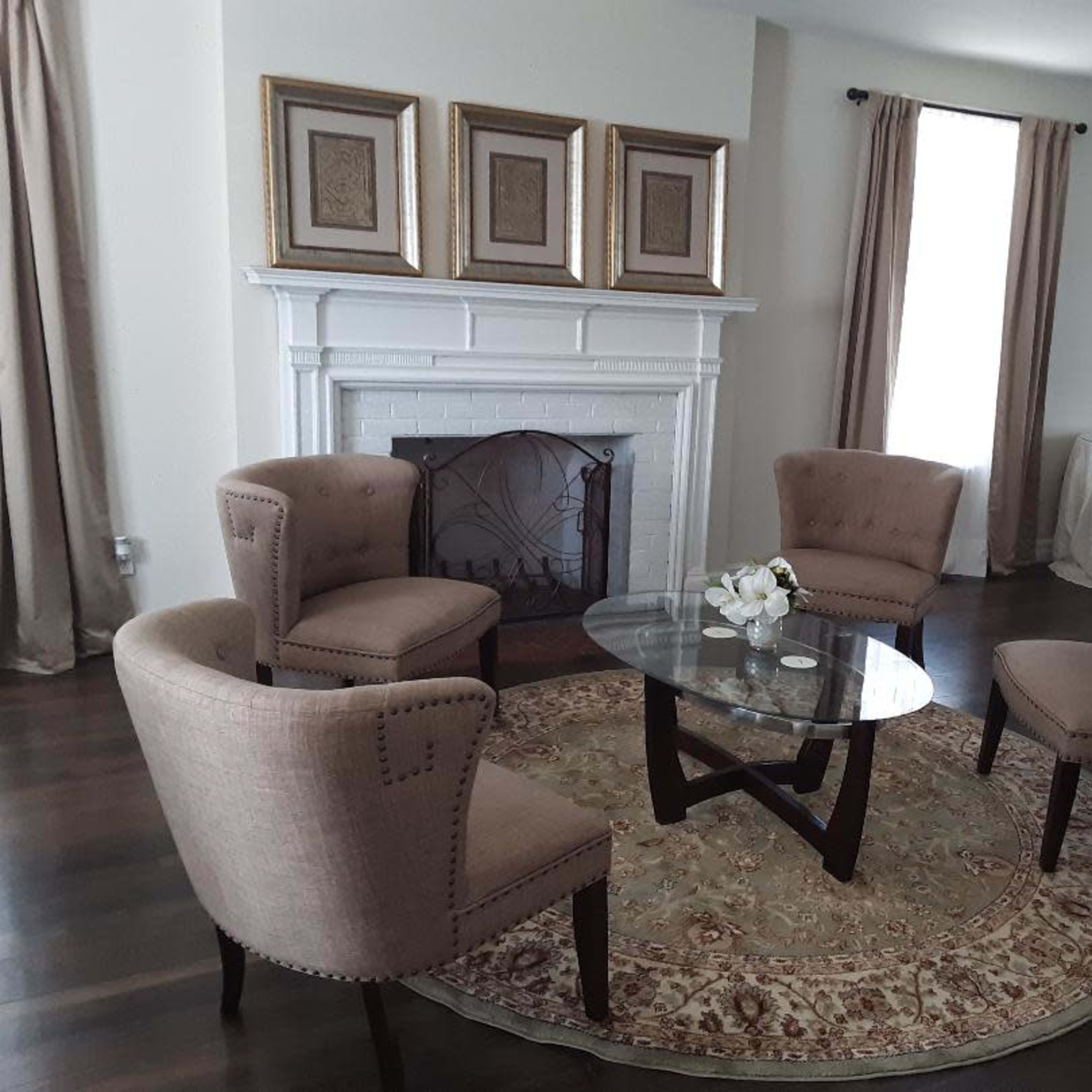 A modern living room with four beige upholstered chairs arranged around a glass coffee table on a patterned rug, in front of a fireplace decorated with three framed artworks.