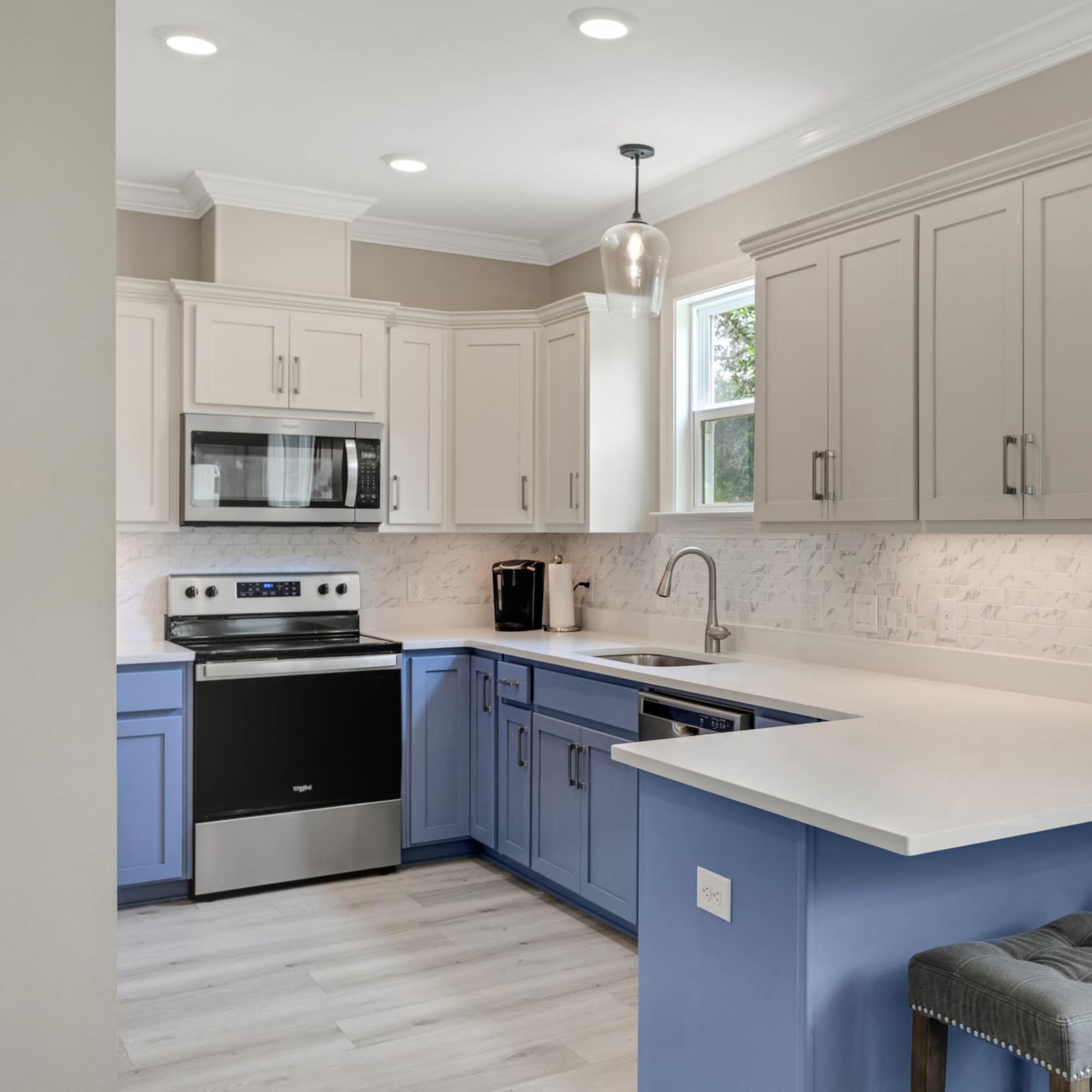 The kitchen features a combination of light gray and blue cabinetry, stainless steel appliances, and a white countertop with two gray stools at the bar.
