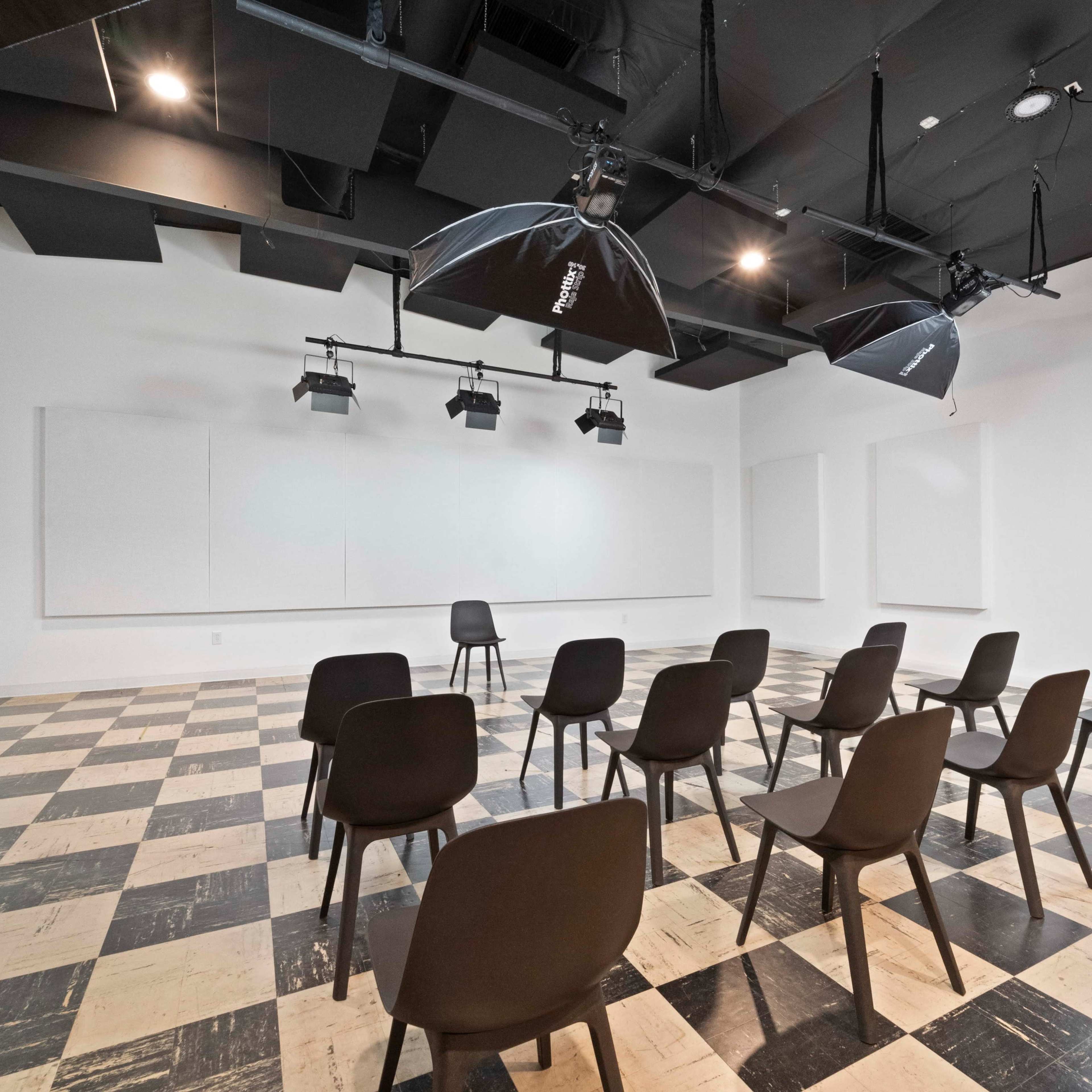 A minimalistic conference room with checkered flooring, black chairs arranged in rows, and overhead lighting fixtures.
