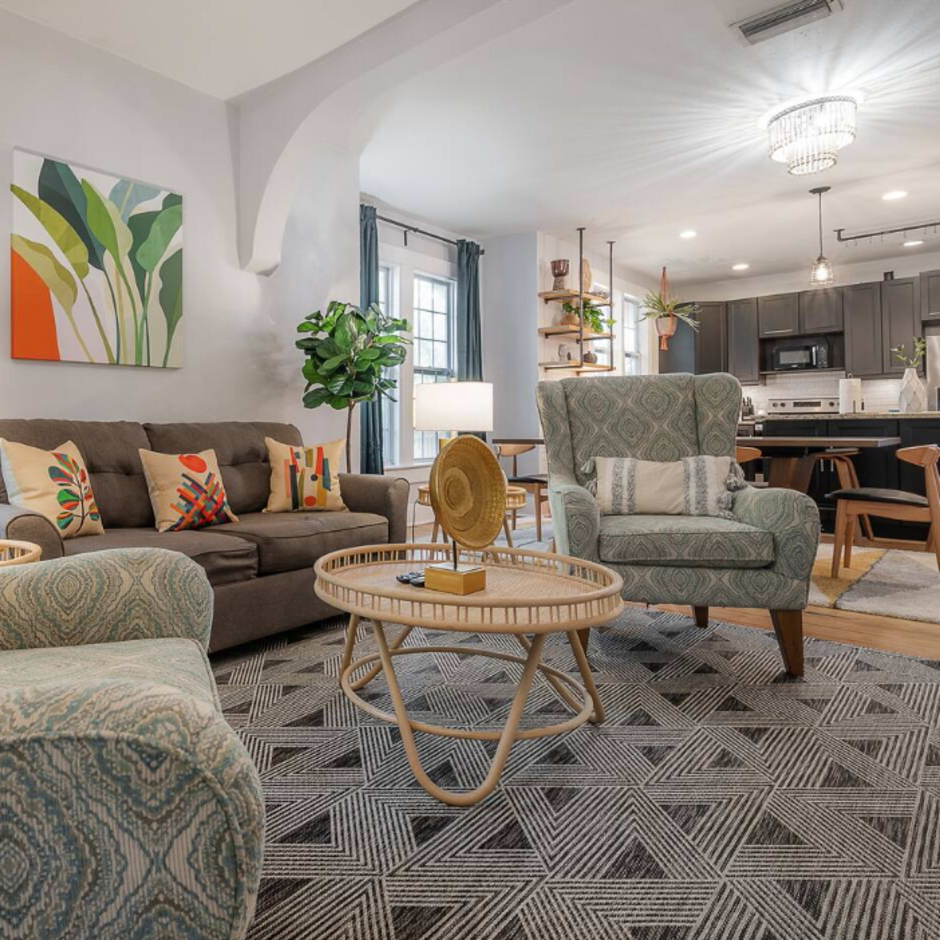 A cozy living room featuring two upholstered chairs, a sofa with decorative pillows, a circular coffee table, and a large plant, with the kitchen in the background.