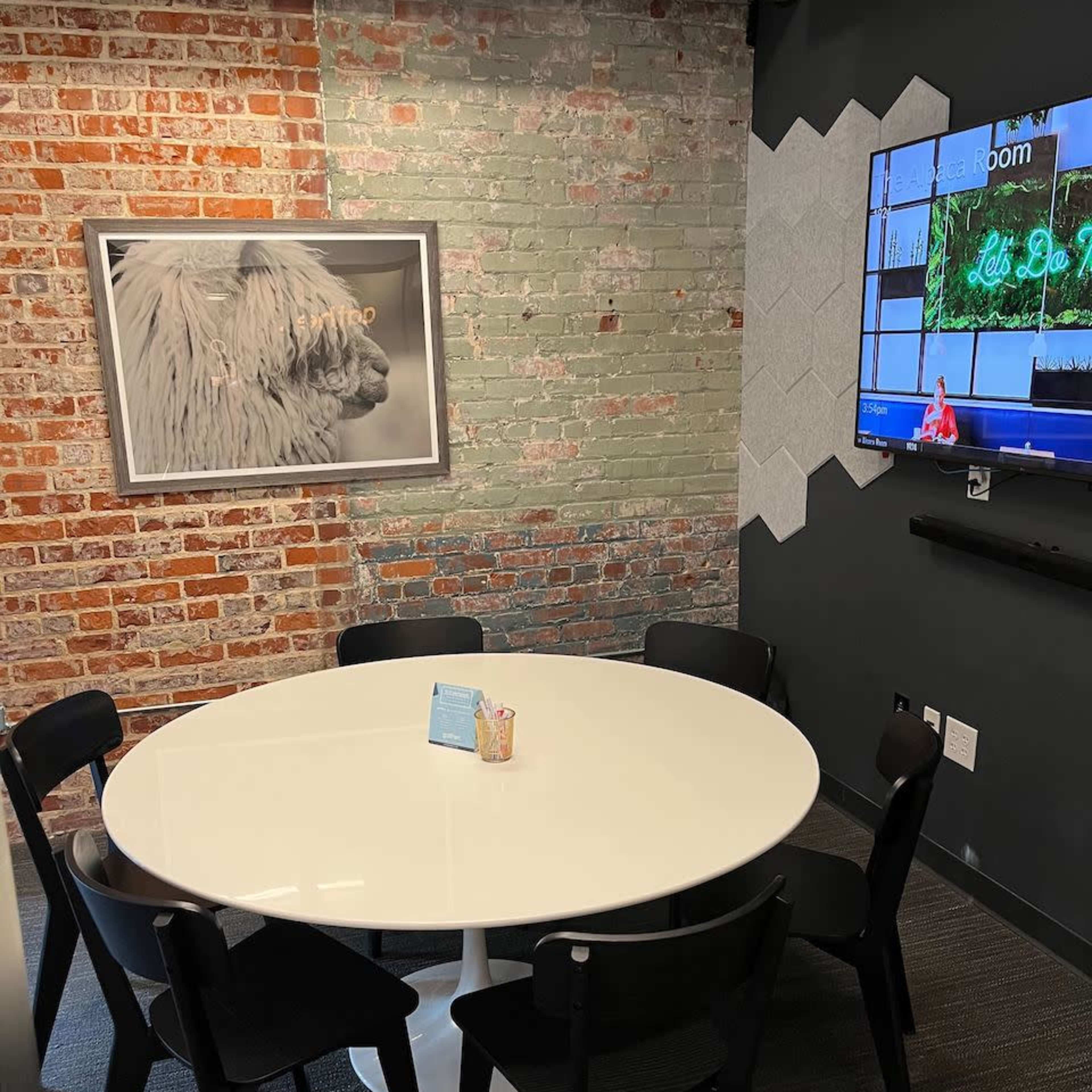 A conference room with a round white table surrounded by black chairs, a brick wall, and a television mounted on a gray wall.