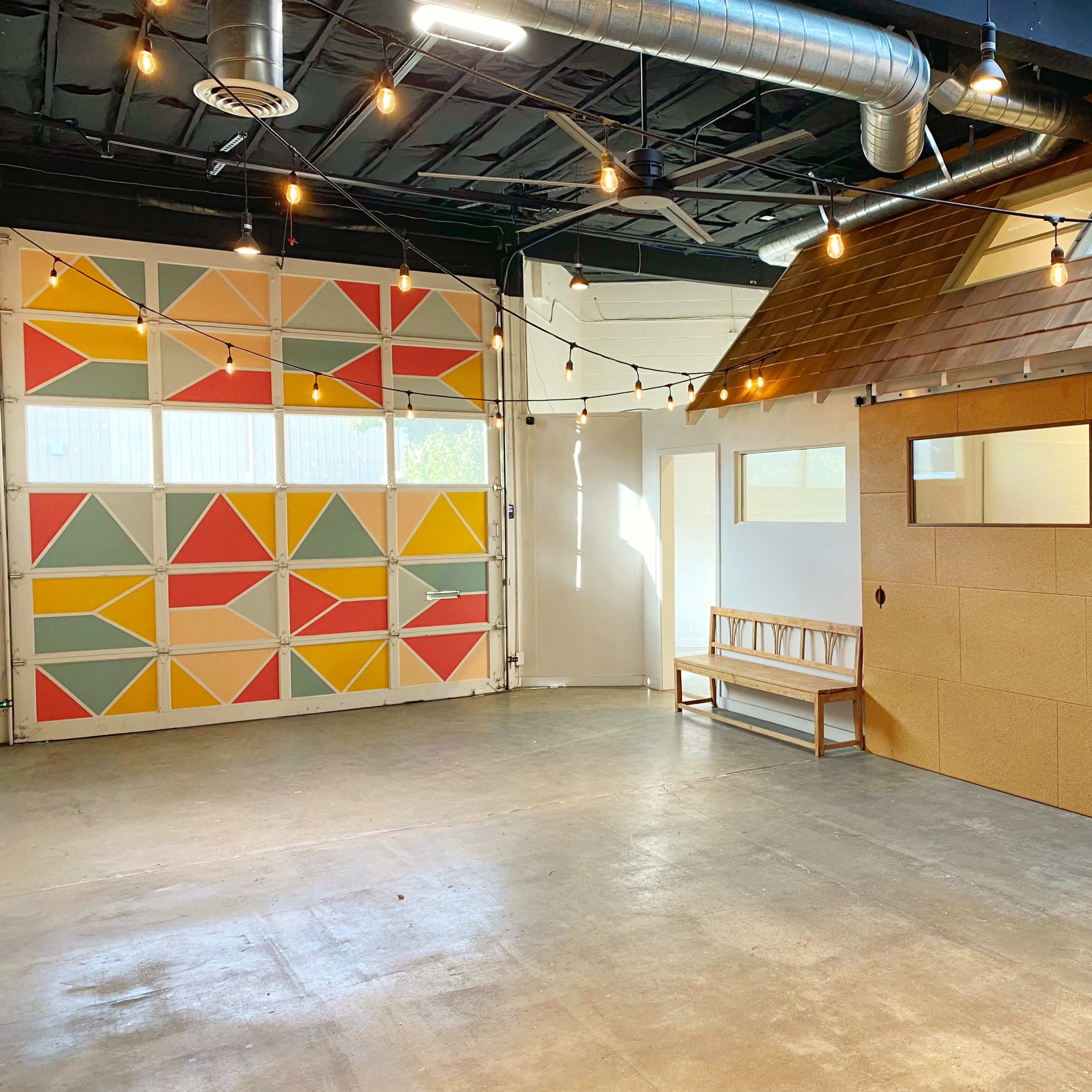 A spacious interior room with a colorful geometric mural on one wall, a wooden bench against another wall, and string lights hanging overhead.