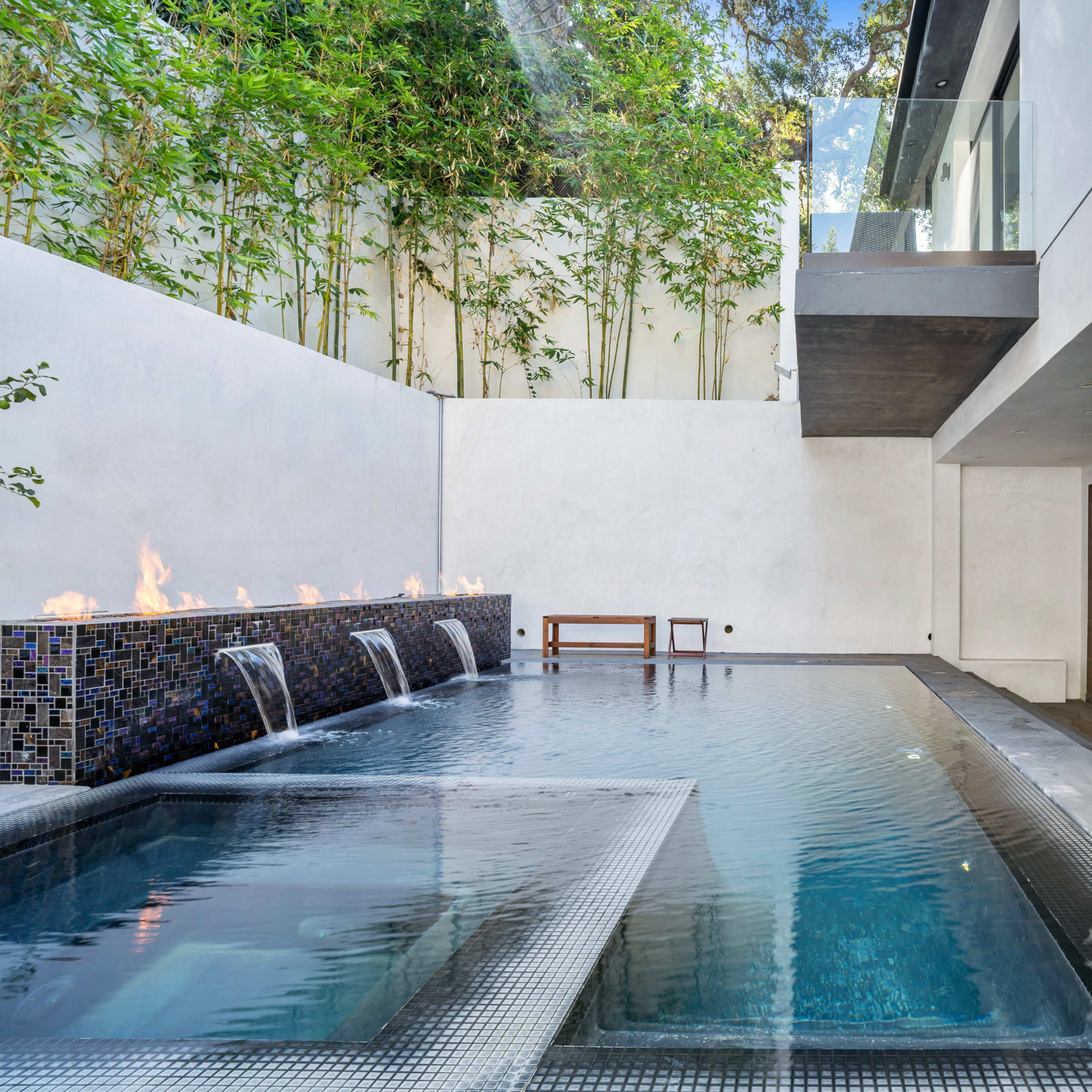 A modern outdoor pool with a water feature and fire elements, surrounded by minimalist architecture and greenery.