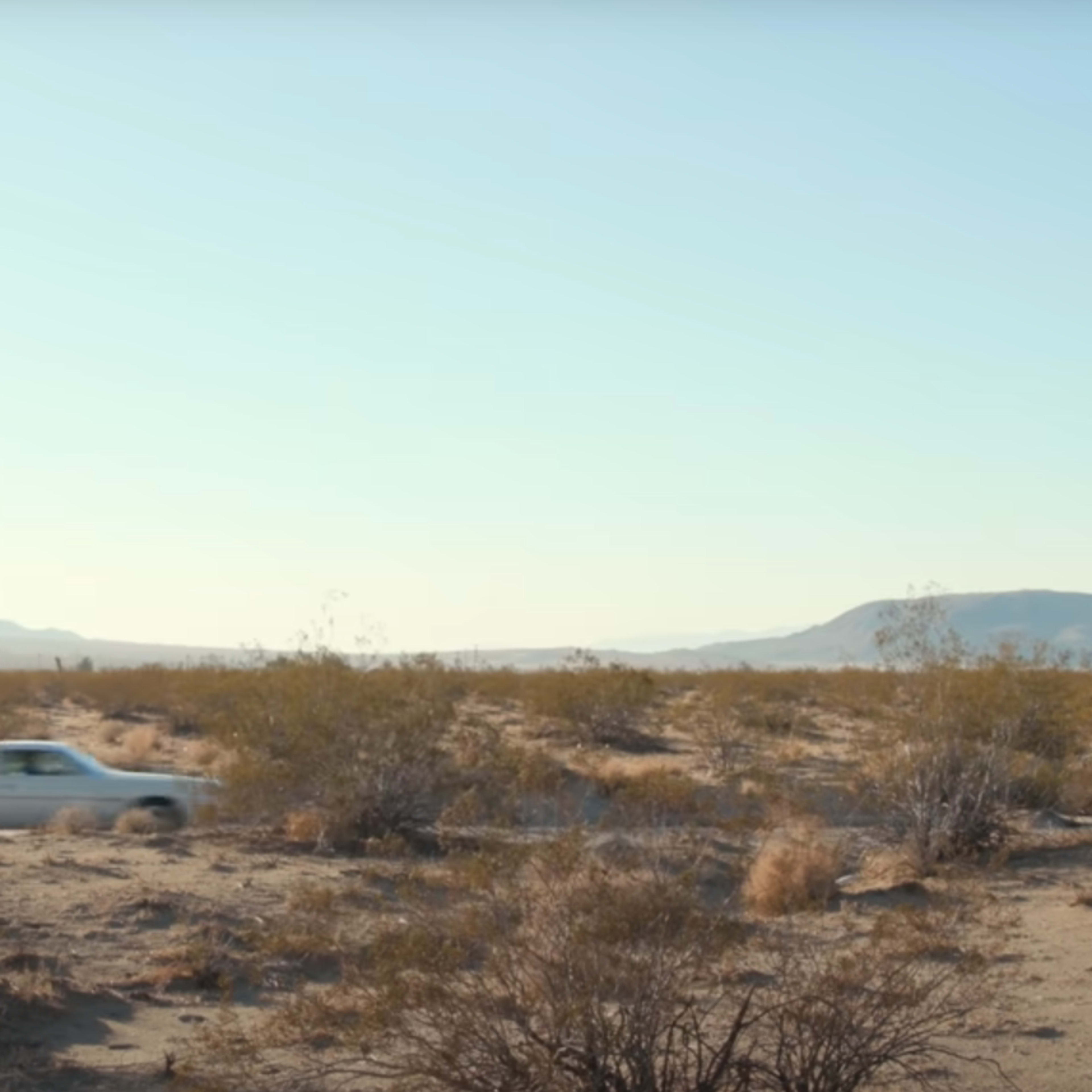 A white car drives through a sandy desert landscape with sparse vegetation and distant mountains.