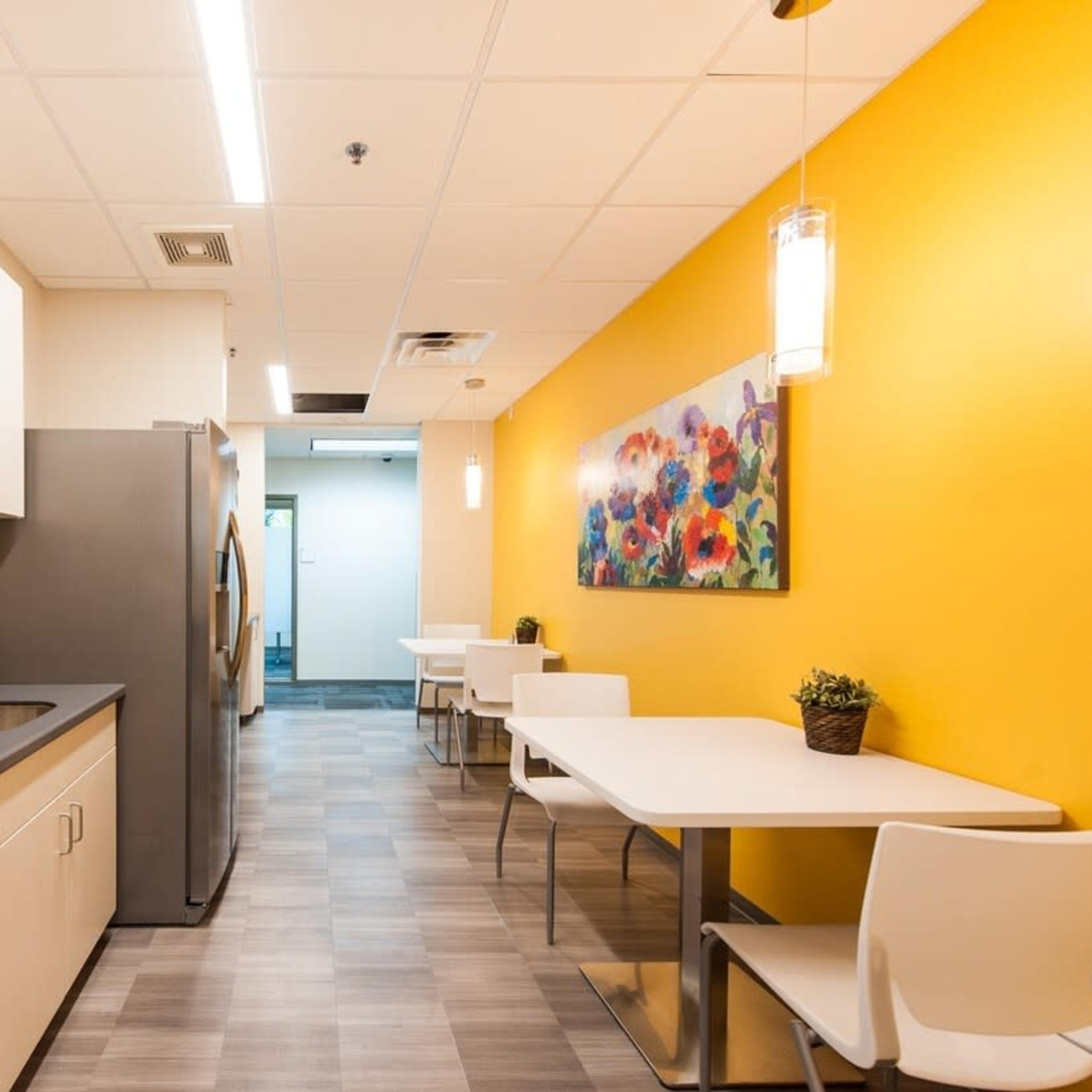A modern break room with a kitchenette on one side, featuring a microwave and a refrigerator, and a dining area with white tables and chairs against a brightly colored wall adorned with a floral painting.