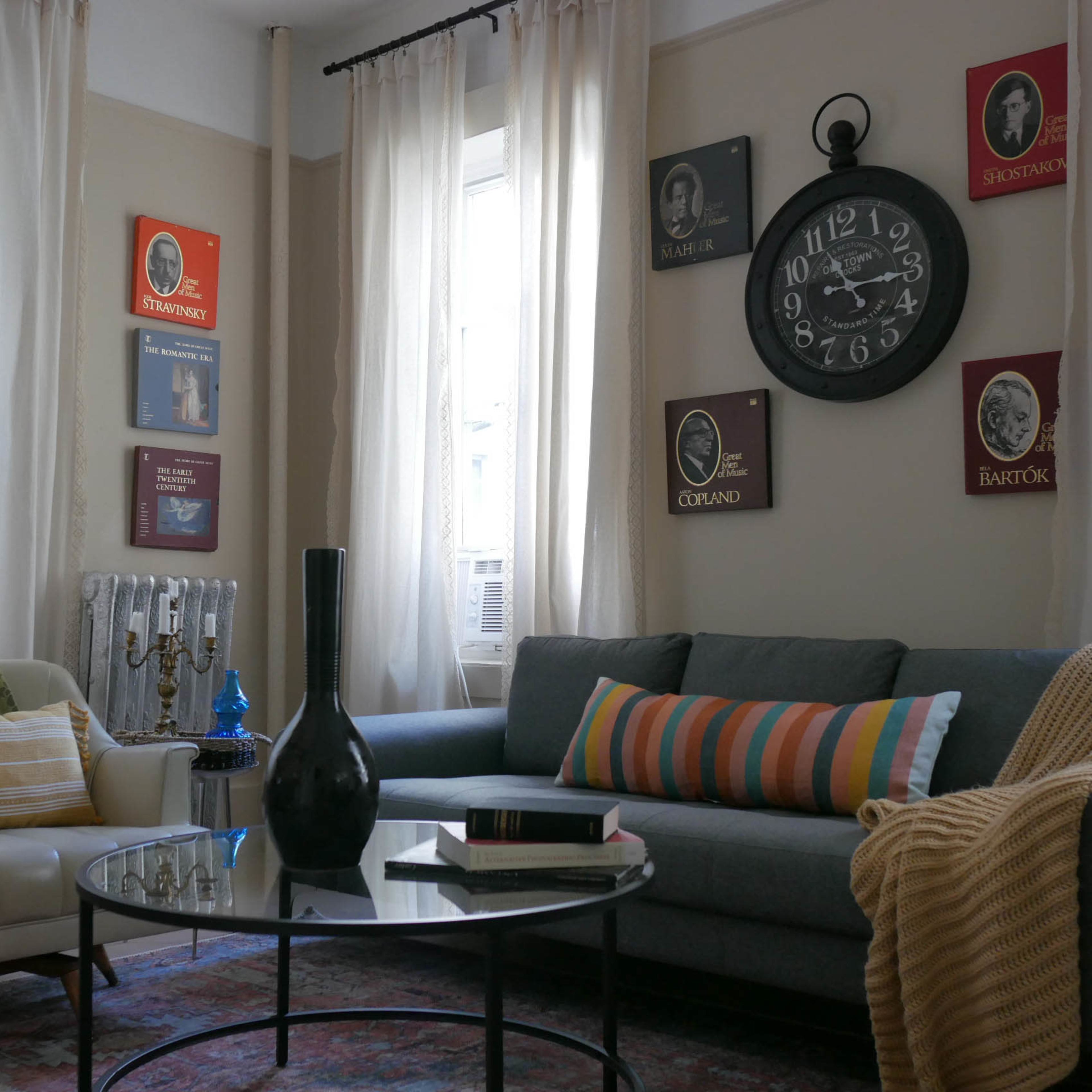 The living room features a gray sofa with colorful cushions, a round glass coffee table, and wall decorations including framed portraits and a large wall clock.