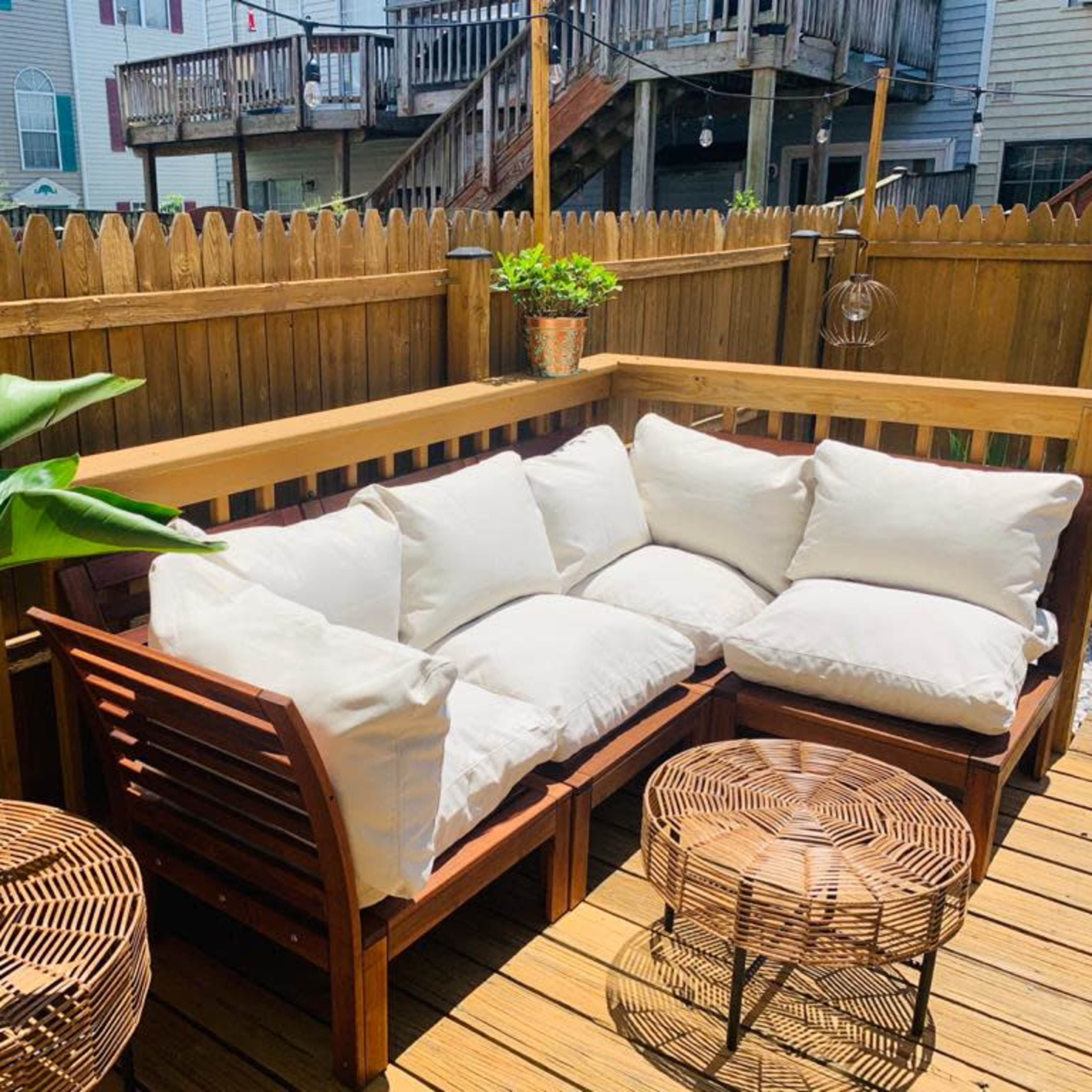 A cozy outdoor seating area with a large cushioned bench, a potted plant, and a woven coffee table, surrounded by wooden fencing.