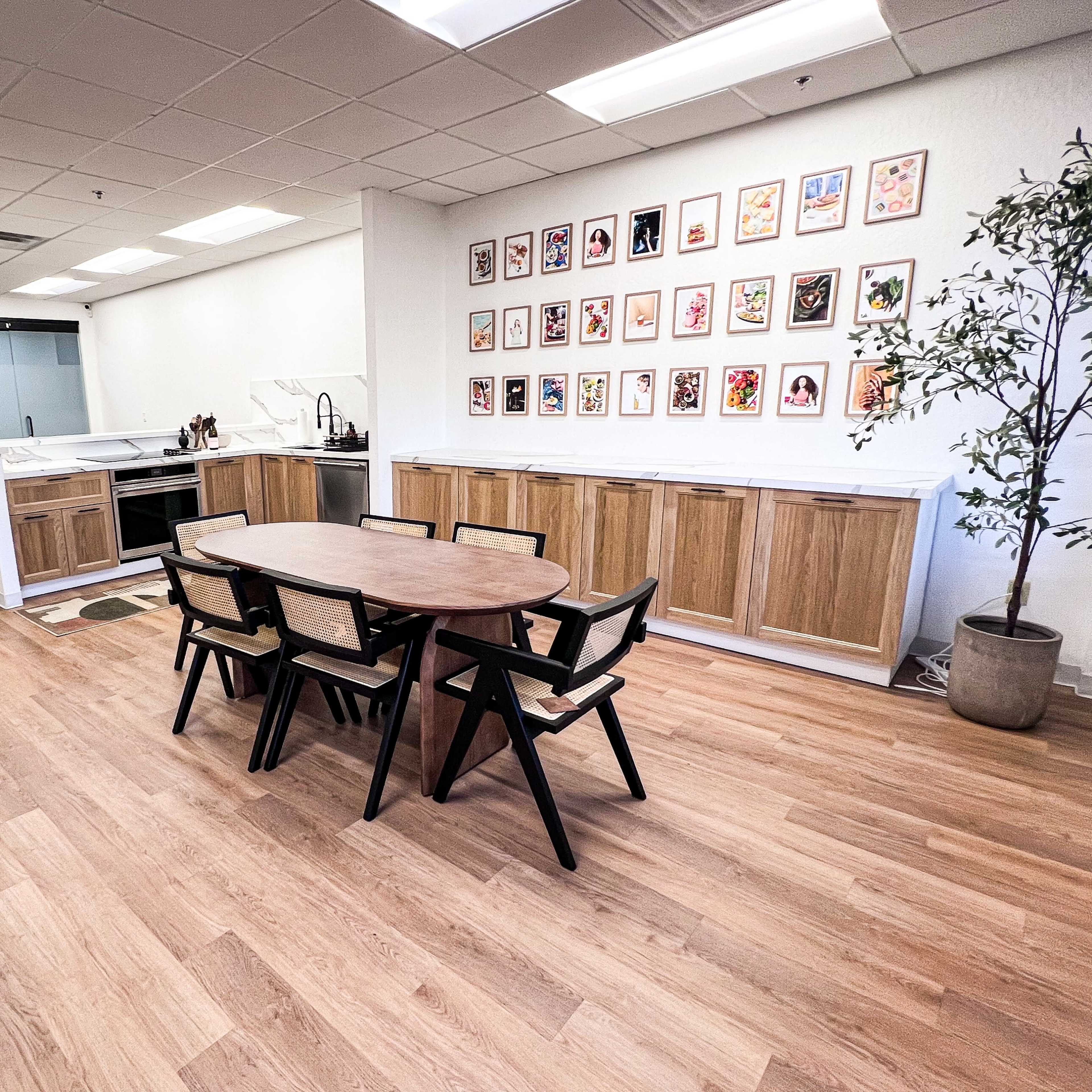 A modern office kitchen area features a wooden dining table surrounded by black chairs, with an art-adorned wall and a kitchen counter in the background.