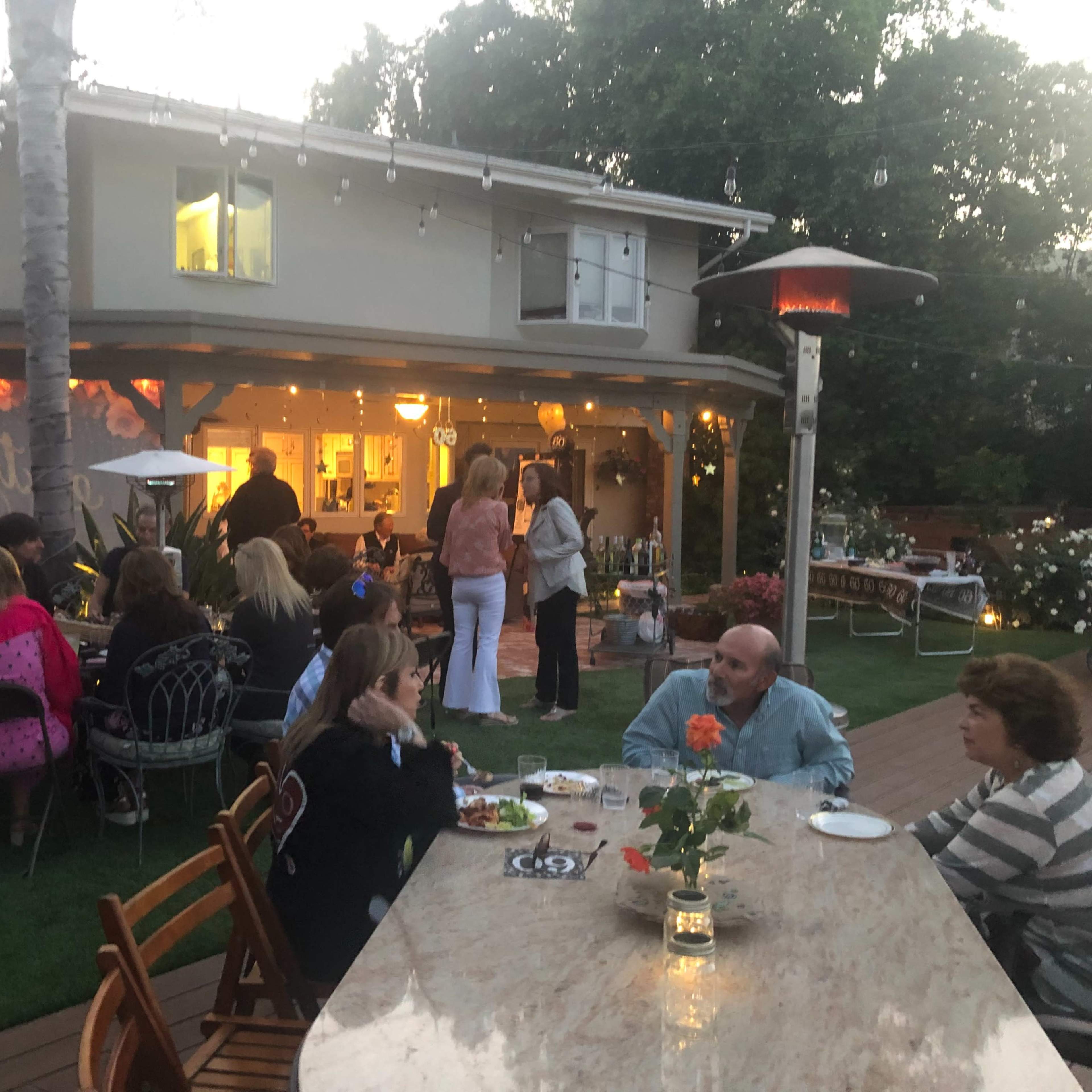A group of people is gathered outside in a backyard, socializing around a long table with food and drinks, while others stand near a house illuminated by warm lights.