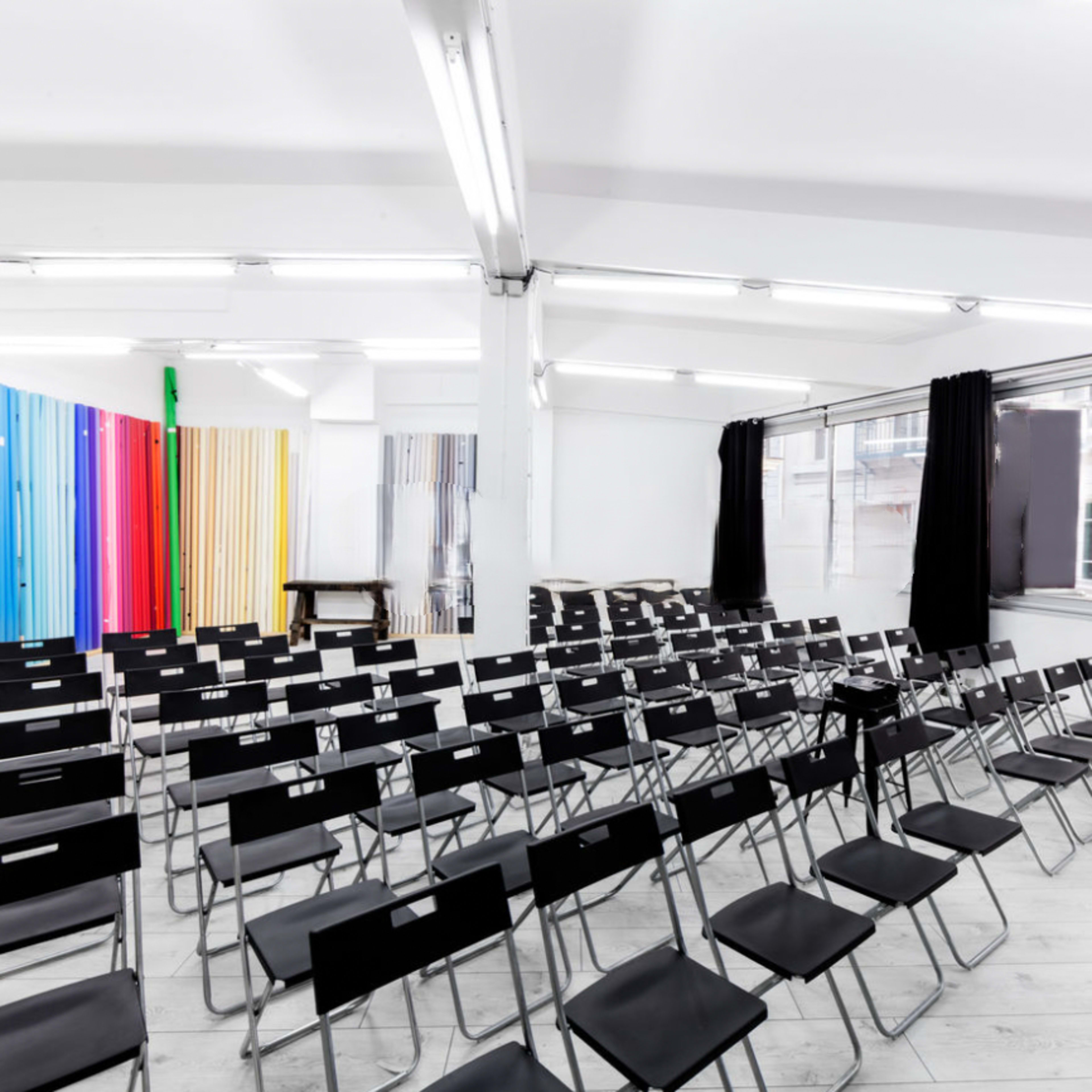 An empty room filled with rows of black folding chairs facing a blank wall, with colorful panels in the background.