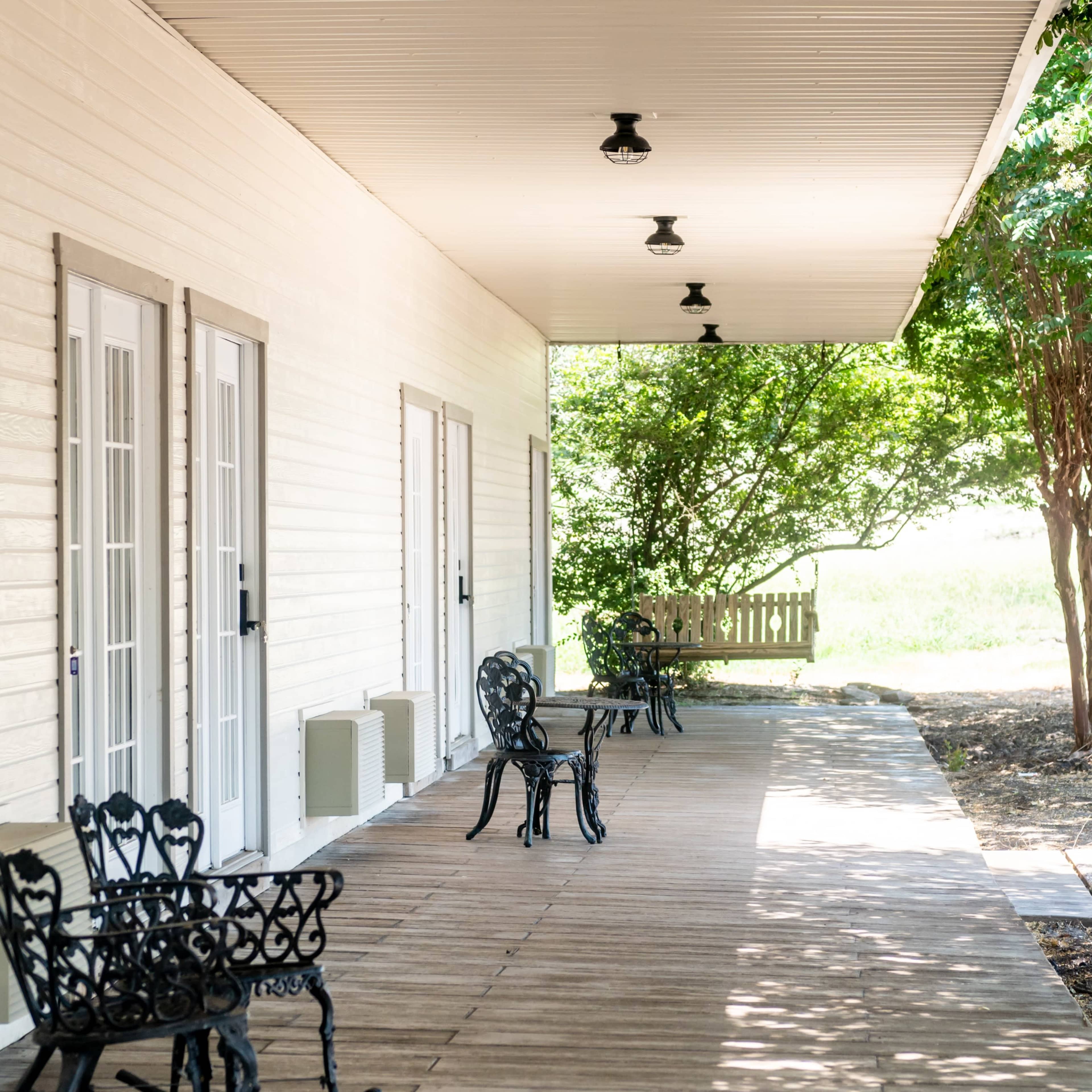 A wooden porch with several black metal chairs arranged along its length, surrounded by greenery and trees.