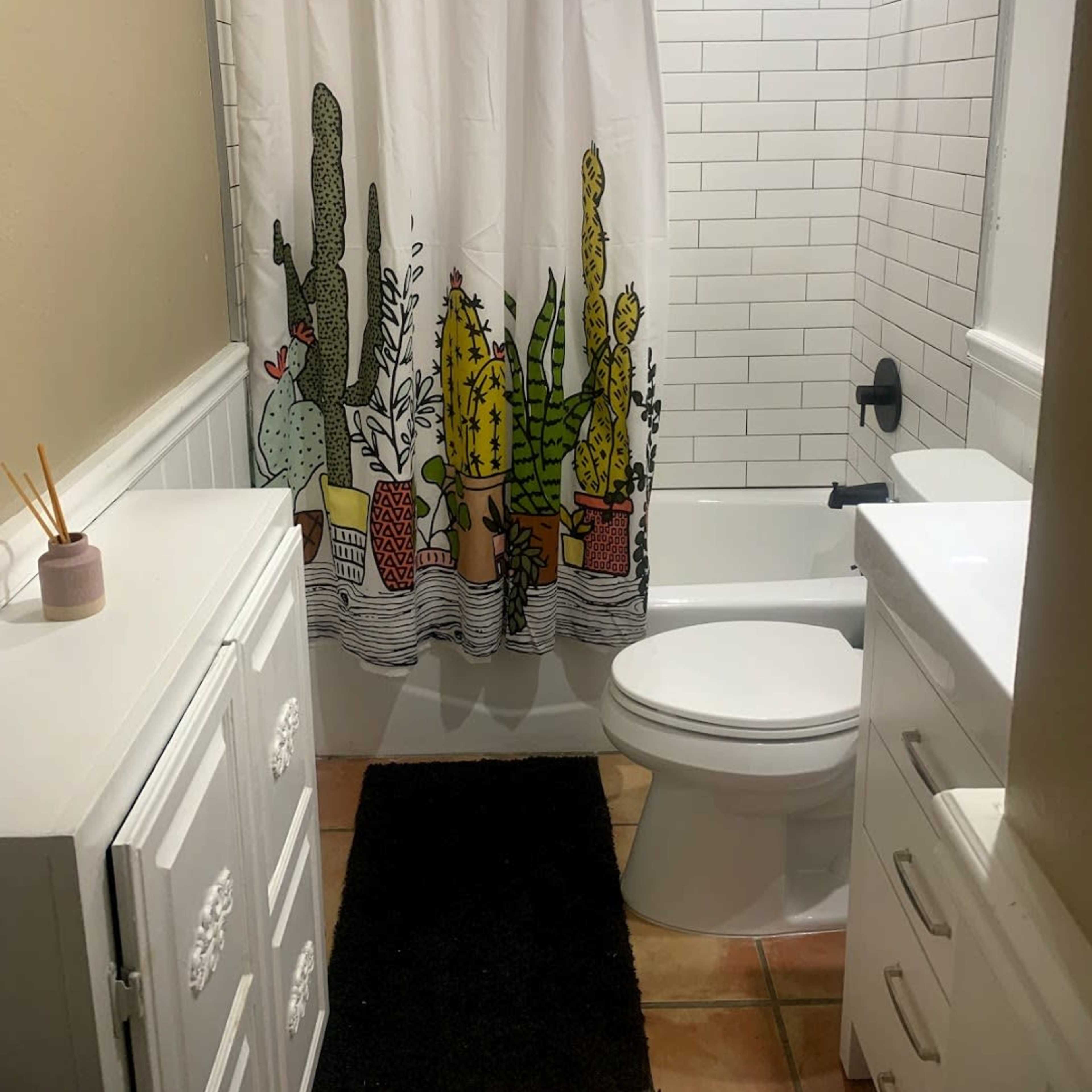 A bathroom features a shower with a cactus-themed curtain, a white vanity, a toilet, and beige tiled floors.