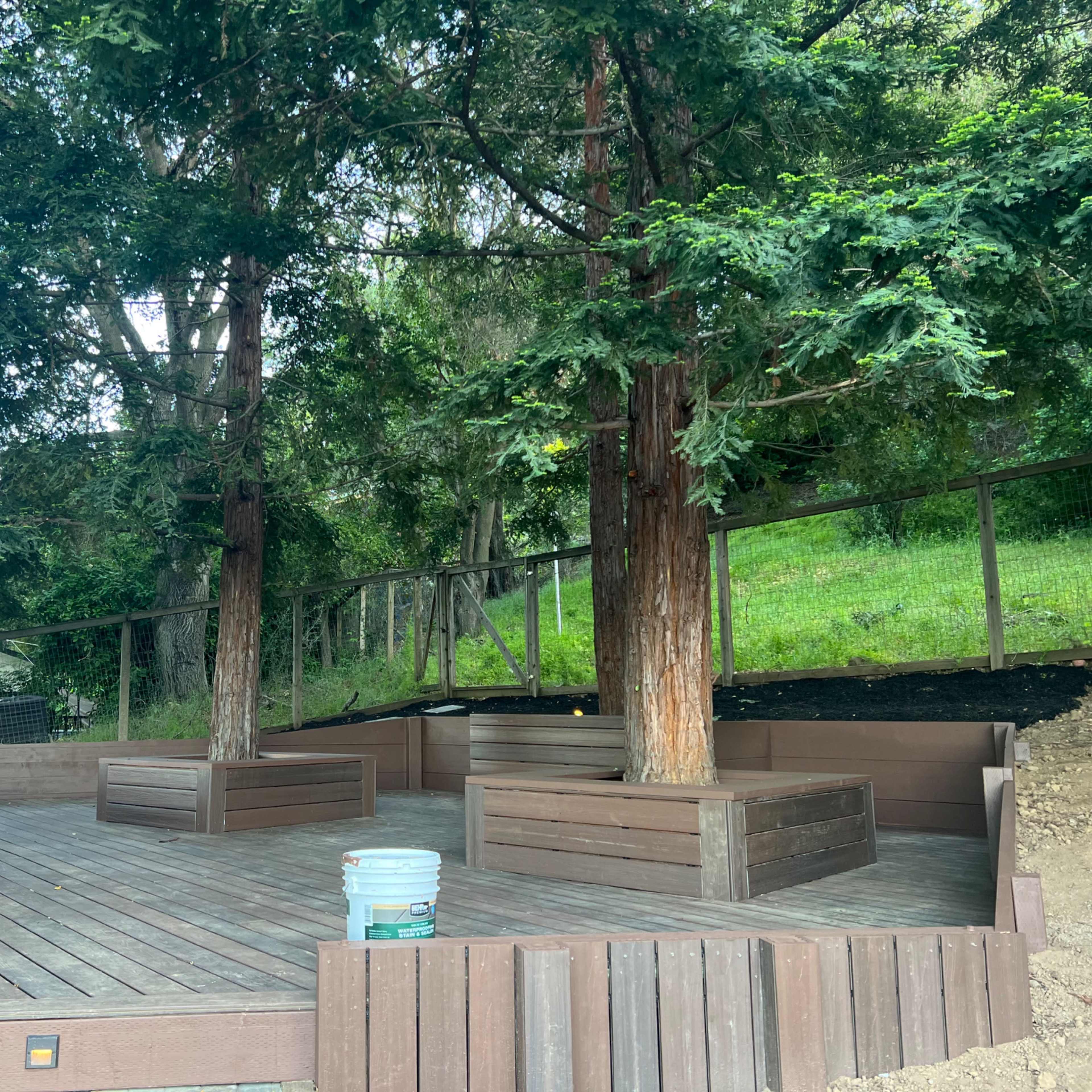 A wooden deck area surrounded by trees, featuring raised planter boxes and a bucket on the ground.