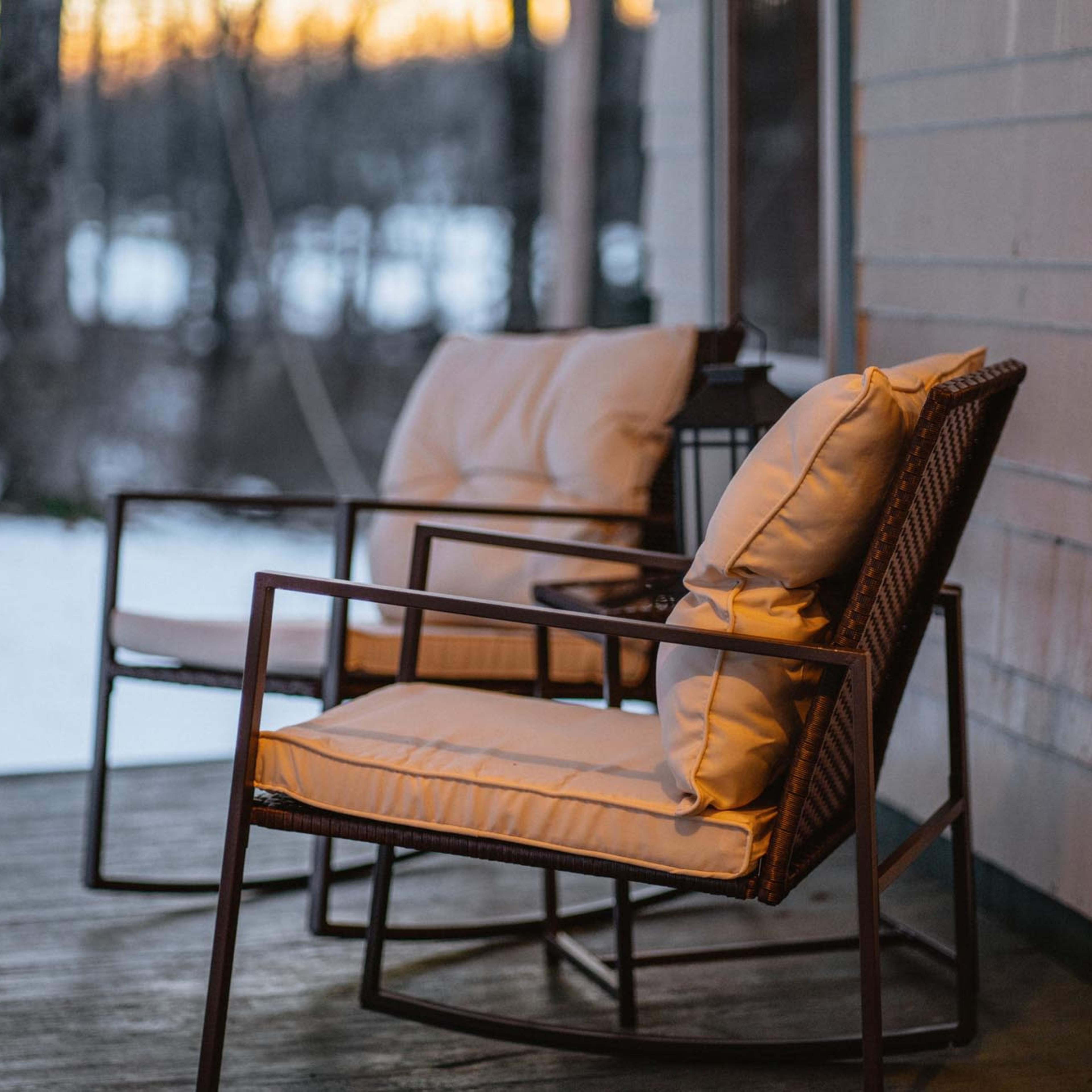Two cushioned rocking chairs sit on a porch overlooking a snowy landscape at sunset.
