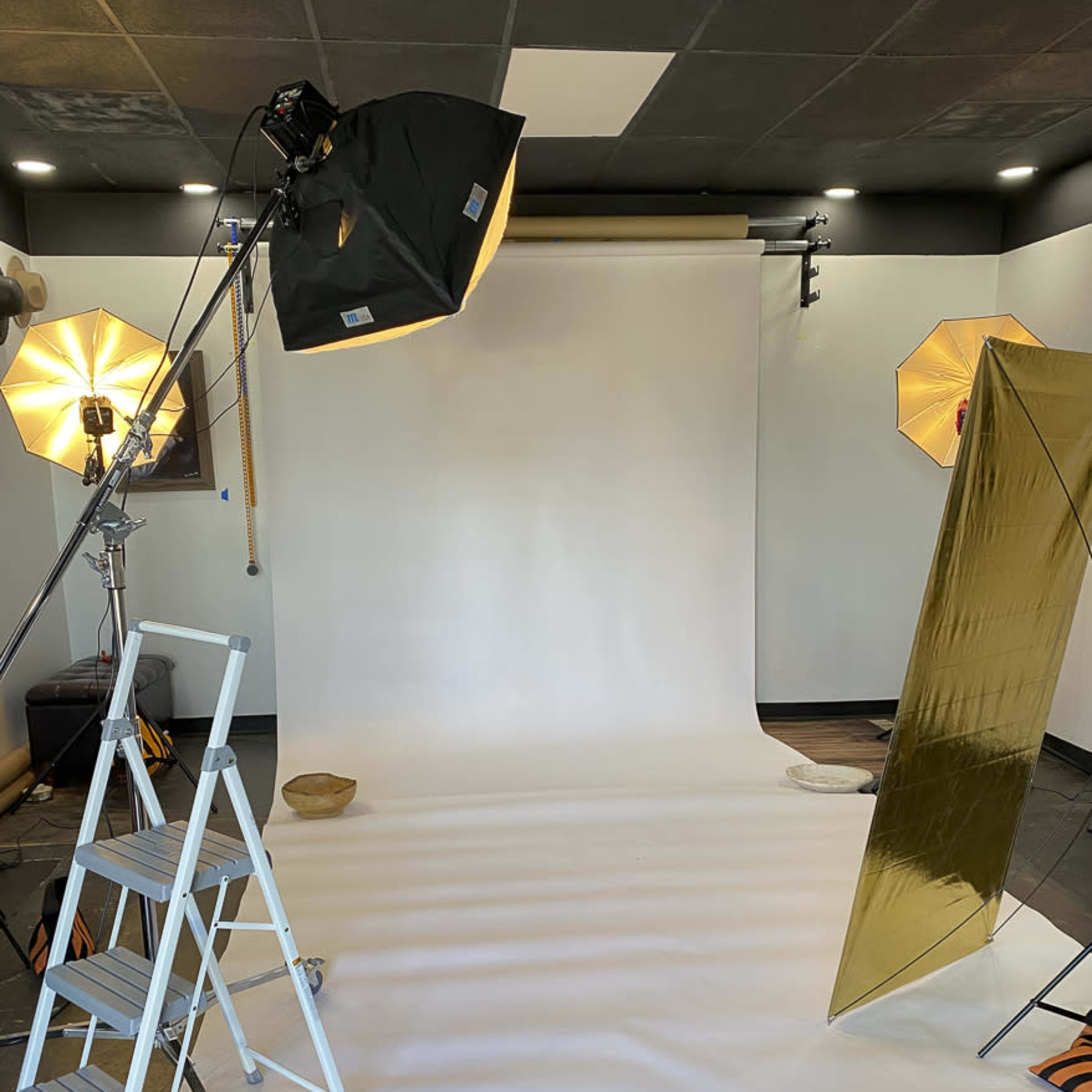 A photography studio setup with a white backdrop, lighting equipment, a ladder, and reflectors arranged around the space.