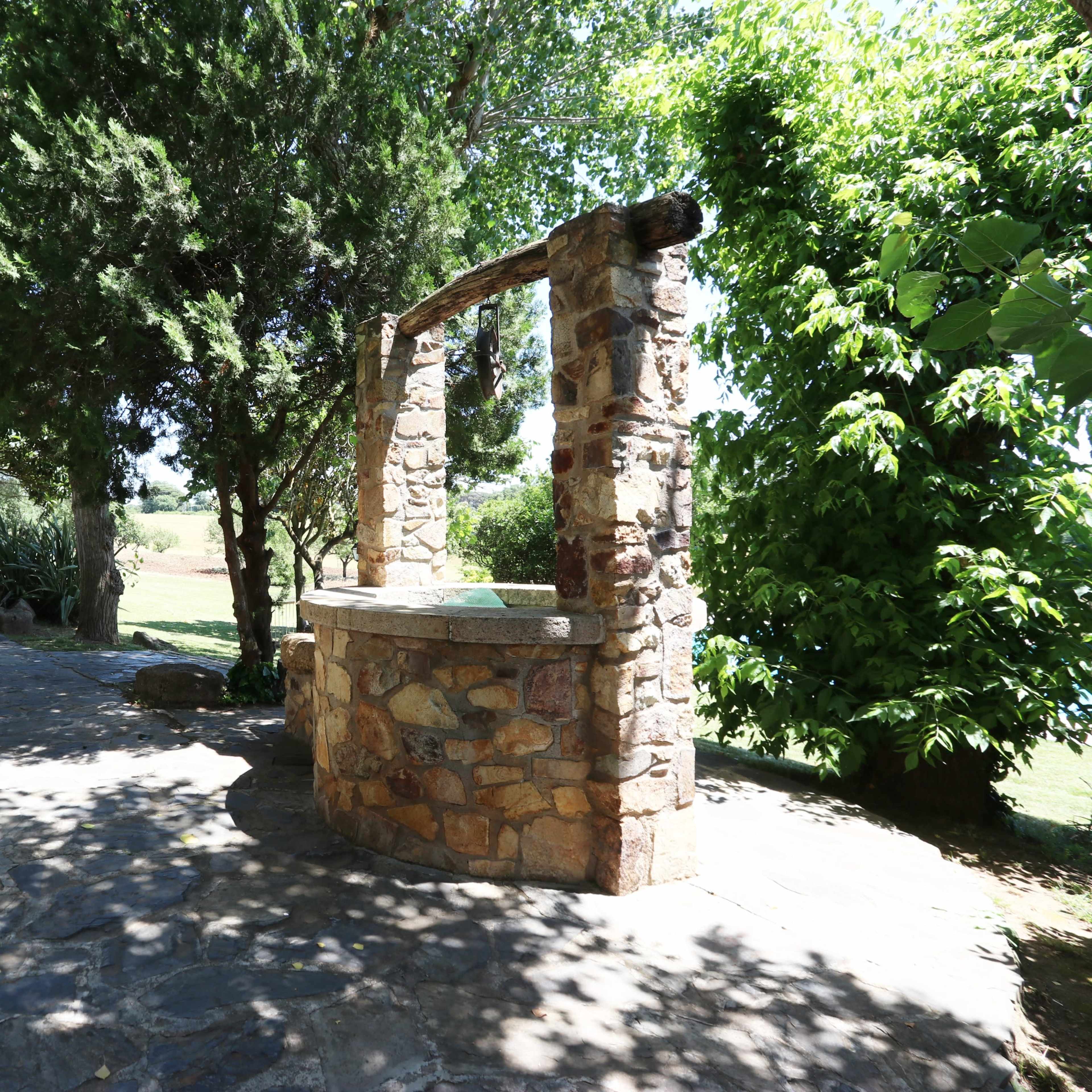 A stone well stands among lush greenery and shaded trees, situated on a paved area.
