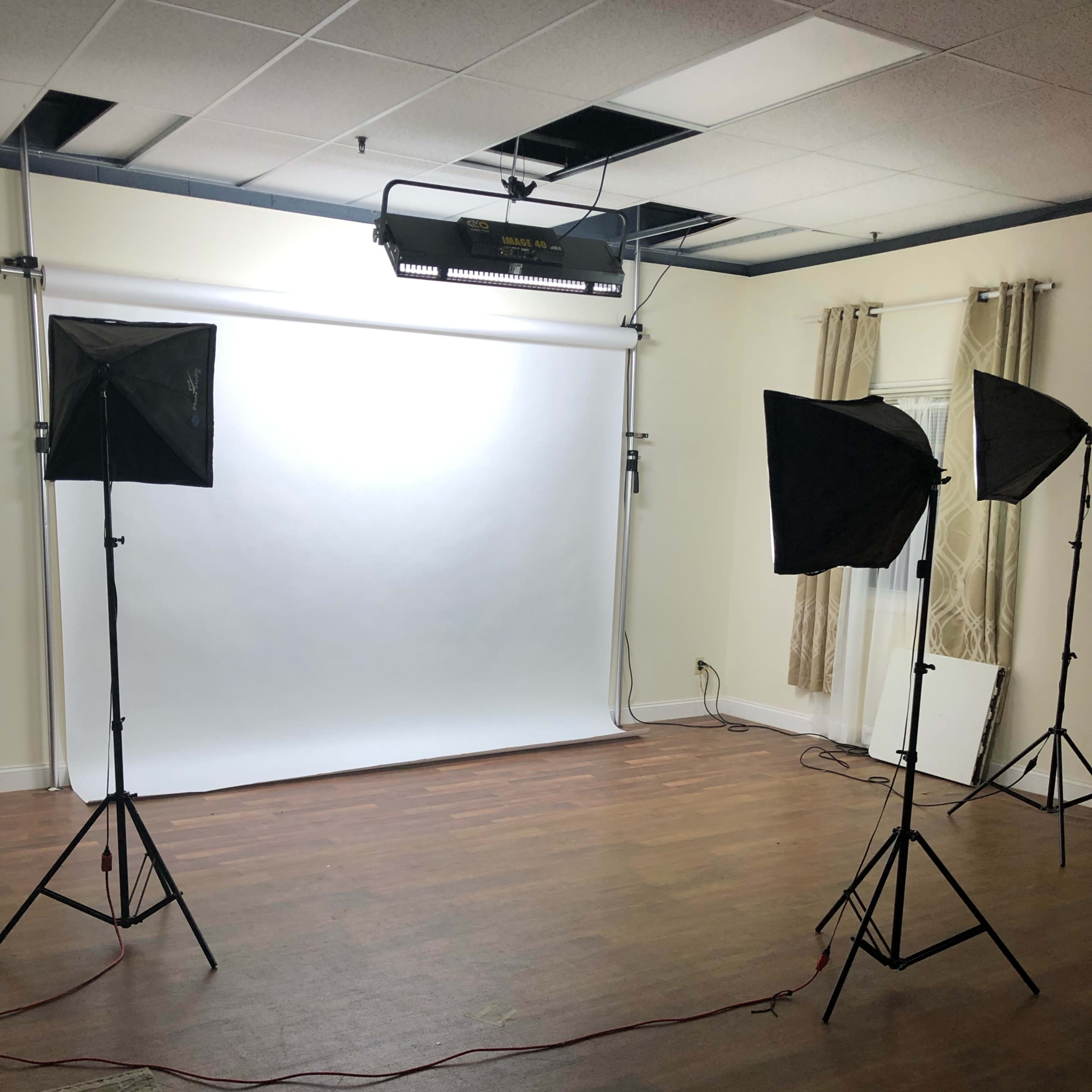 A photography studio setup with a white backdrop, three softbox lights, and a wooden floor.
