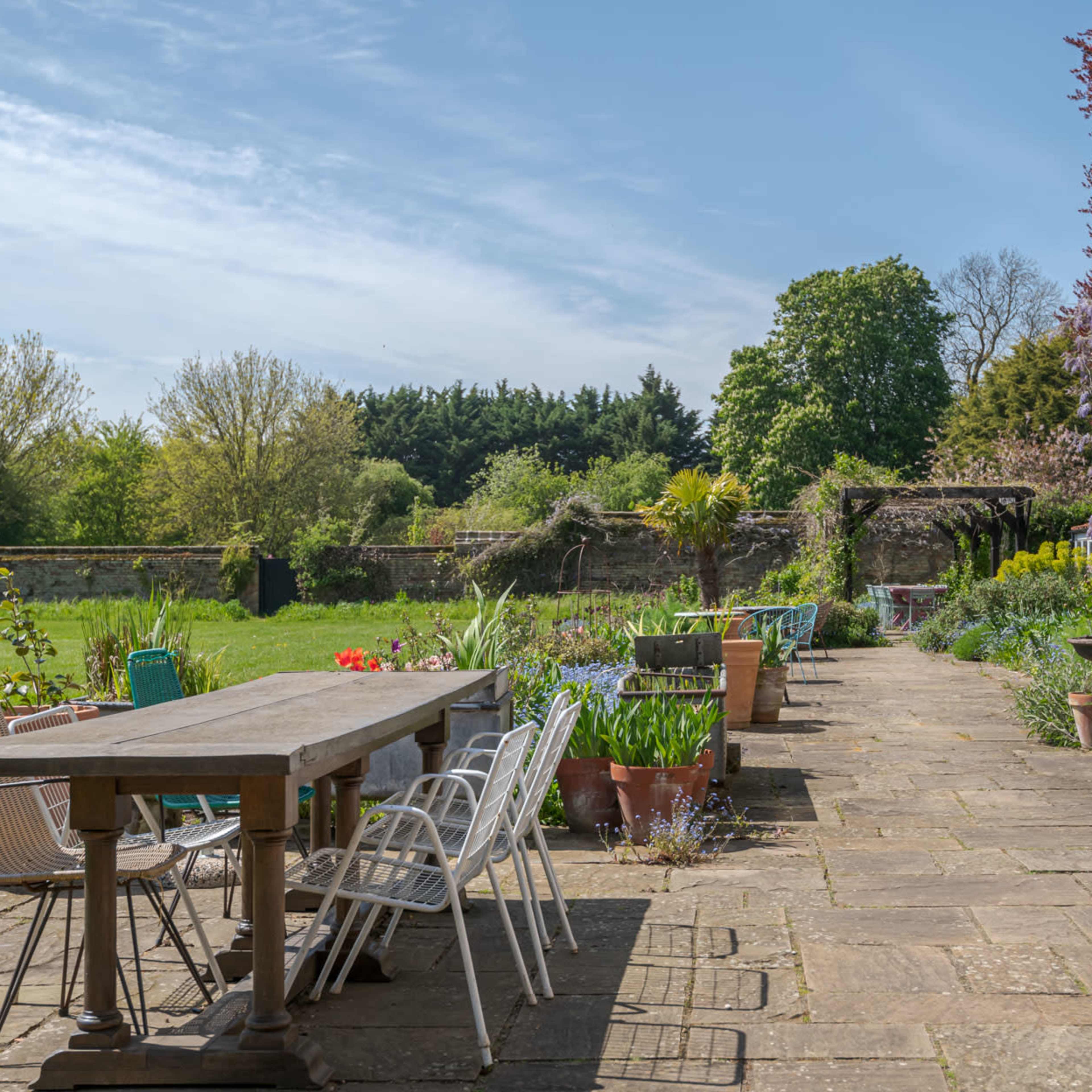 A stone patio with a long wooden table and various potted plants stretches alongside a grassy area and a stone wall.