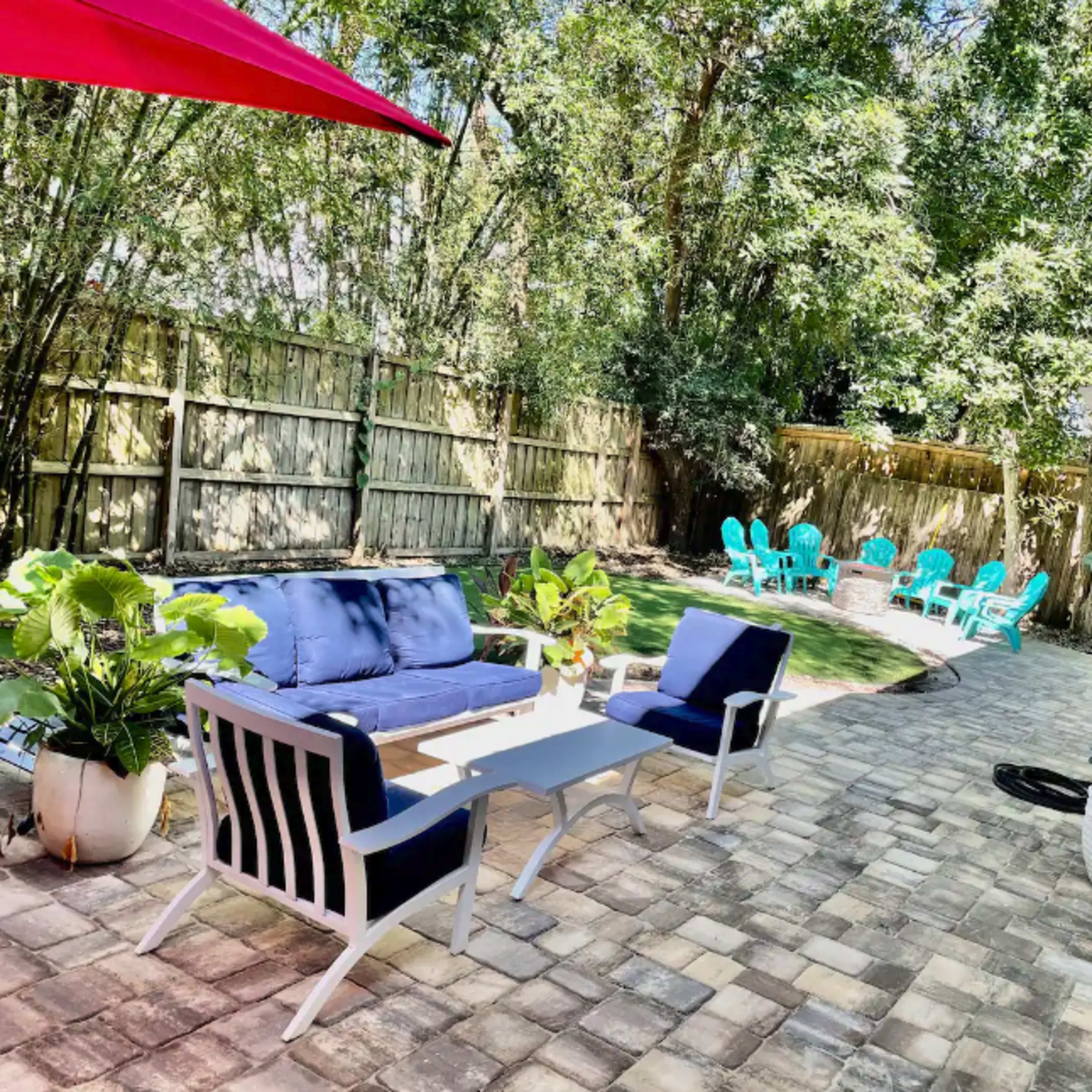 A backyard patio with a blue sectional sofa, a glass coffee table, and several turquoise lounge chairs arranged on a stone-paved surface.