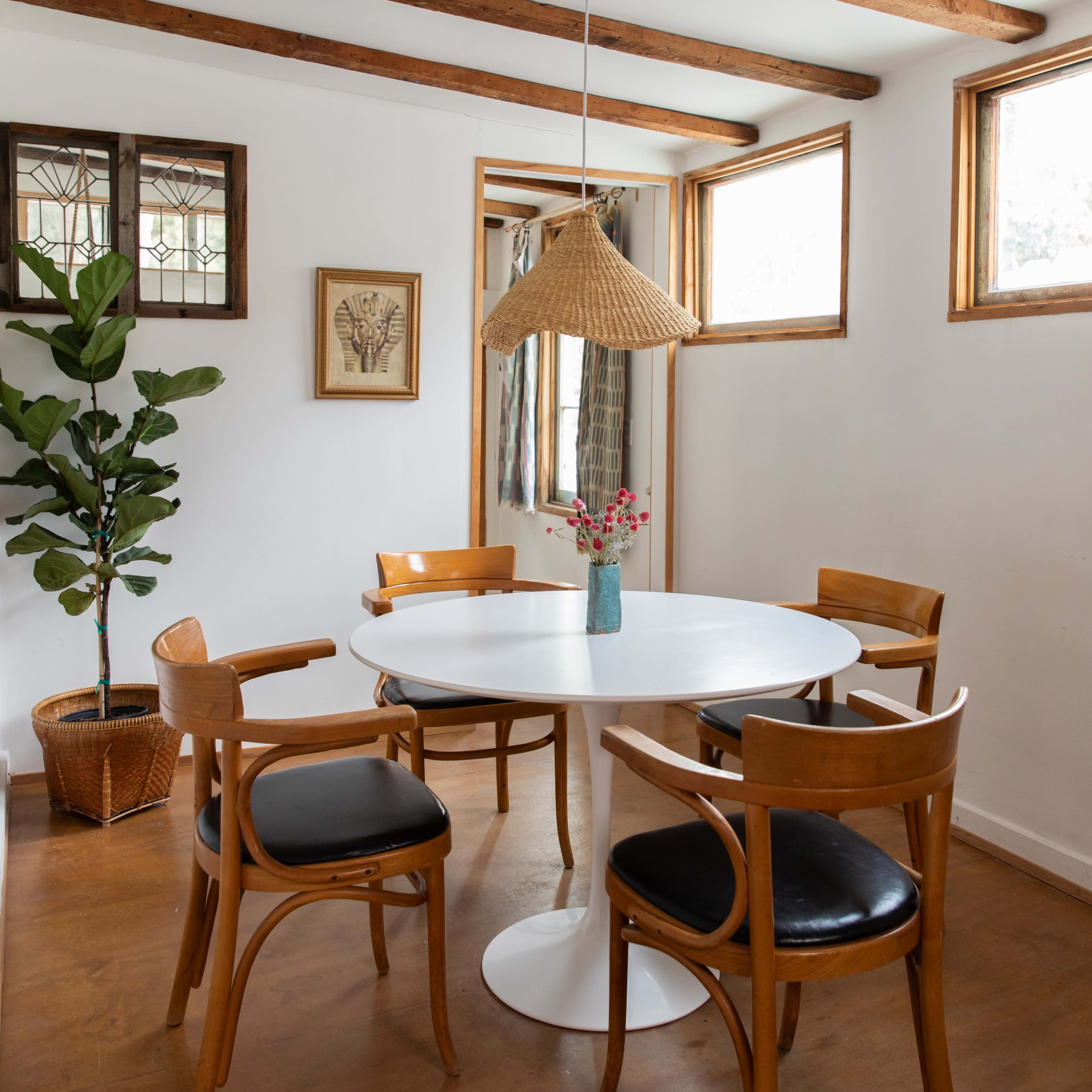 A dining area with a round white table surrounded by four wooden chairs, a plant in a basket, and natural light coming through the windows.