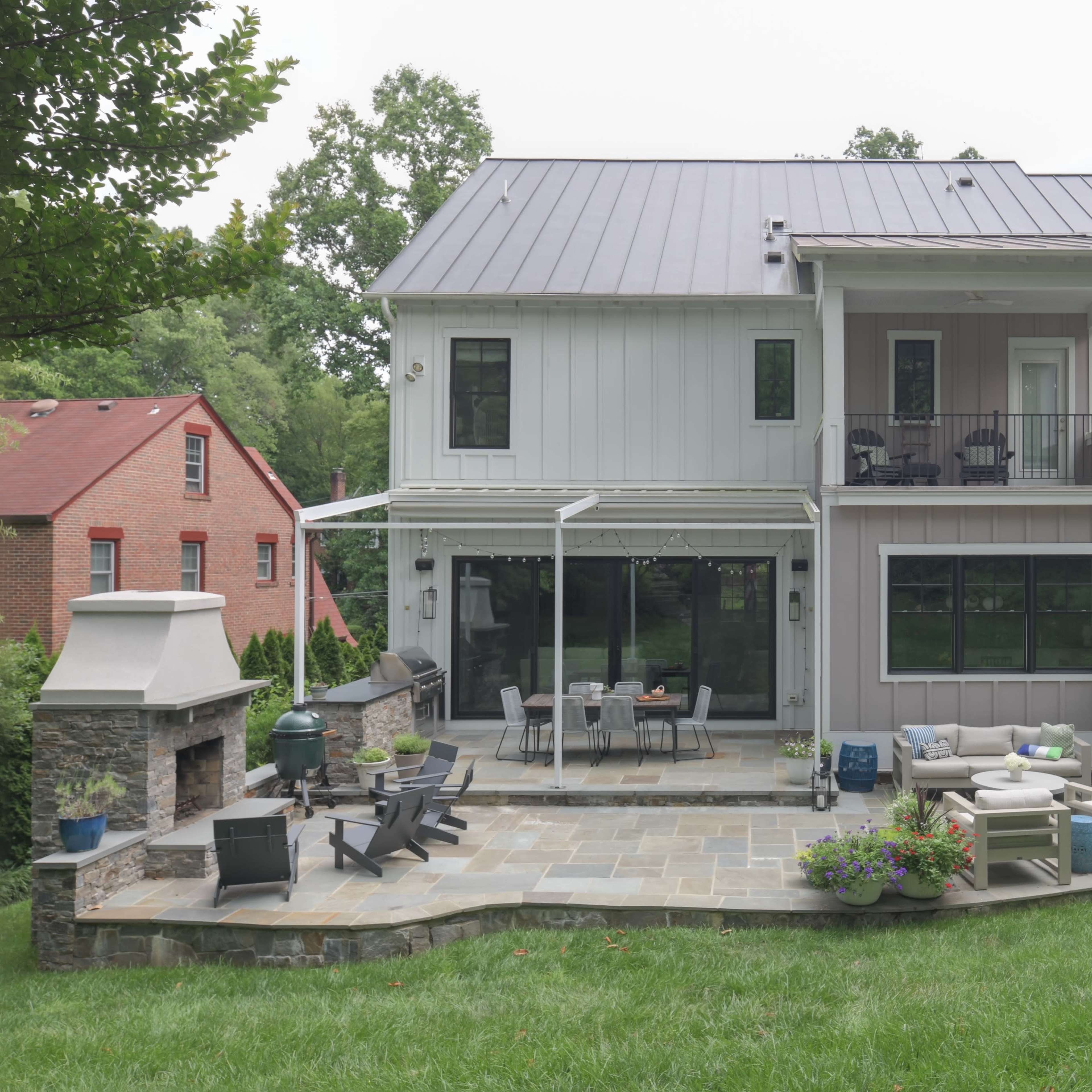A modern two-story house with a metal roof, large windows, and a landscaped backyard featuring a stone patio and outdoor seating.