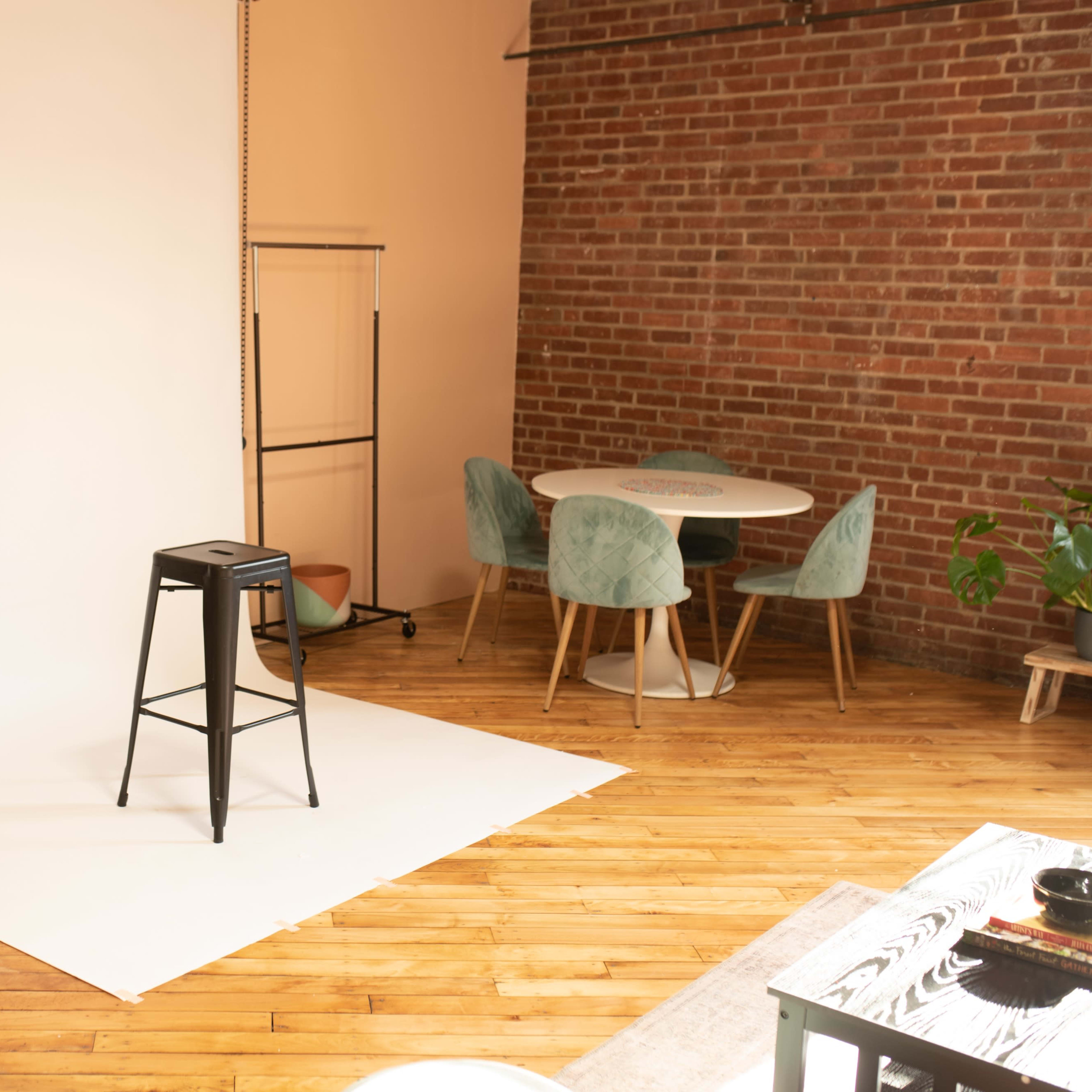 A studio space with a black stool on a white backdrop, surrounded by a wood floor, a round table with chairs, and a tall tripod.