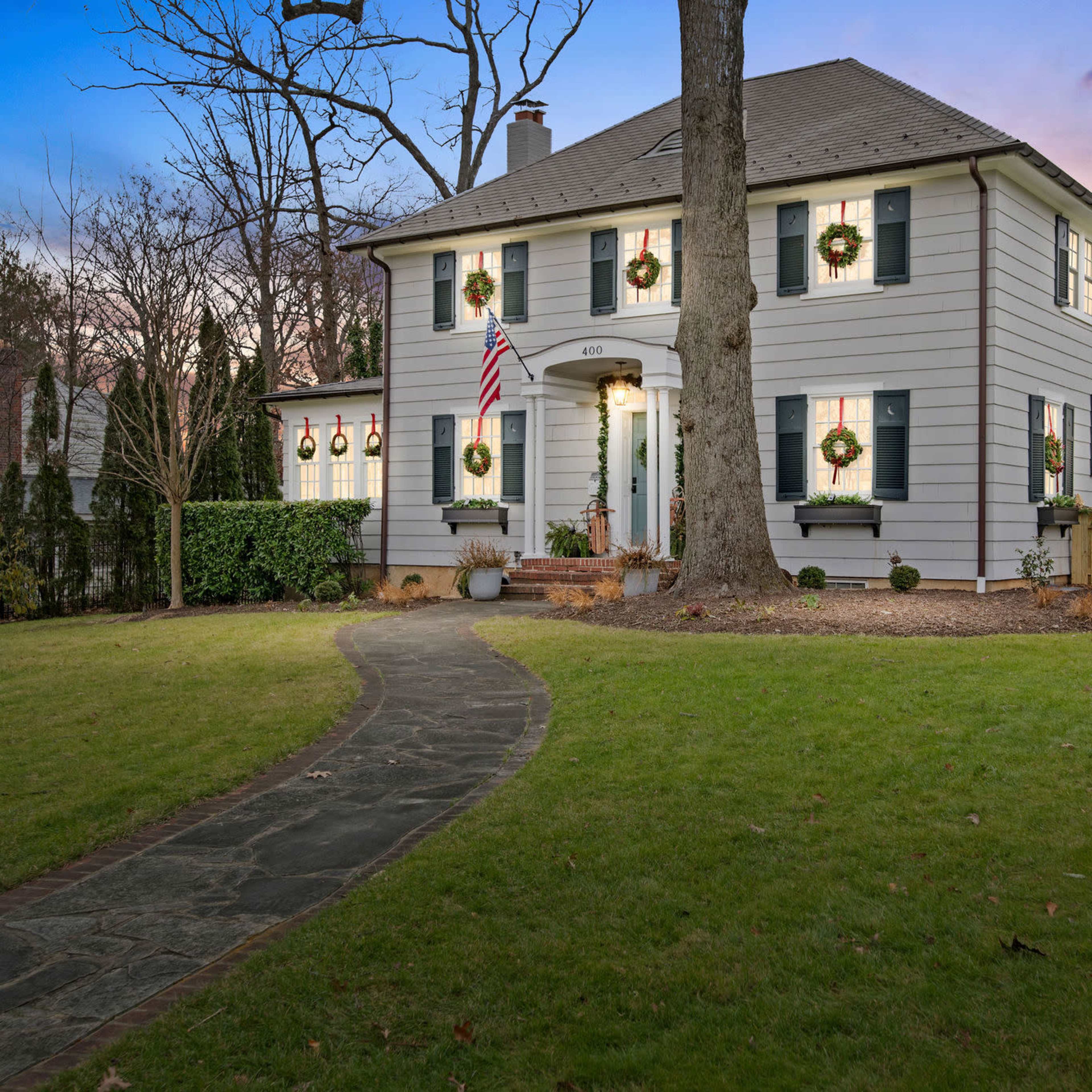 A two-story house with white siding and a front path lined with greenery stands decorated with wreaths and an American flag, set against a colorful sunset.