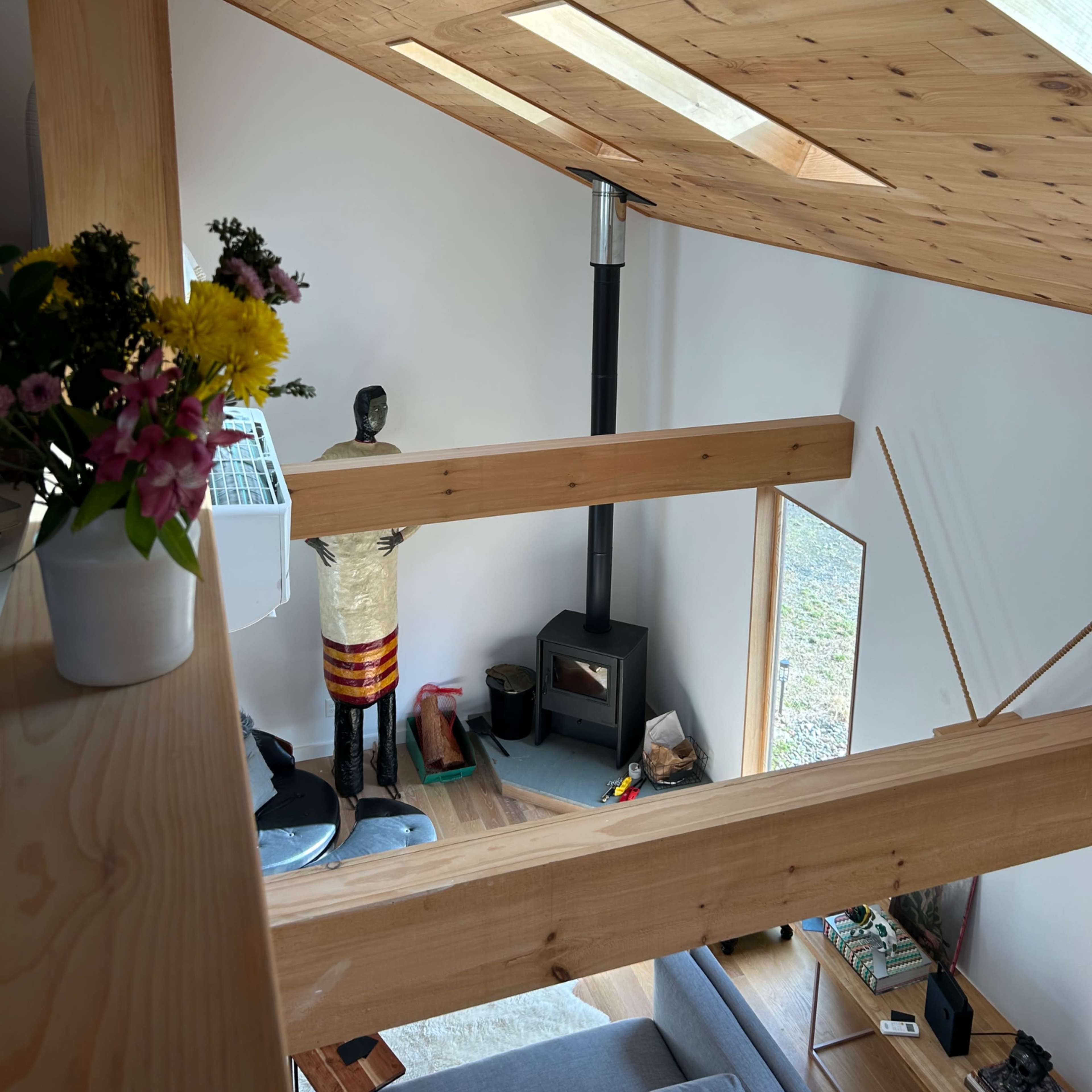 The interior of a modern wooden cabin with a sloped ceiling, featuring a wood stove and a balcony view of the living space below.