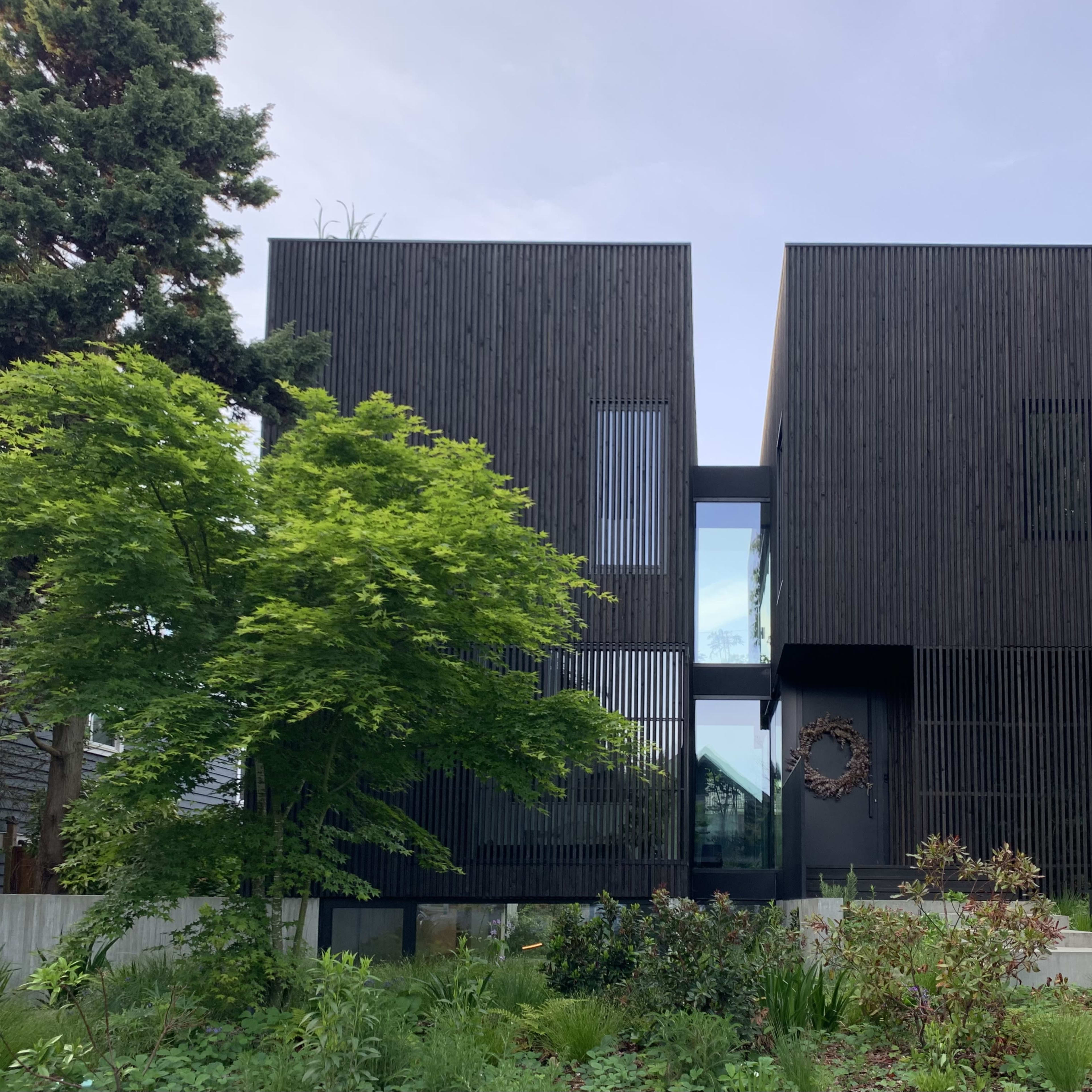 A modern black wooden house with two distinct rectangular sections, flanked by greenery and a landscaped garden.