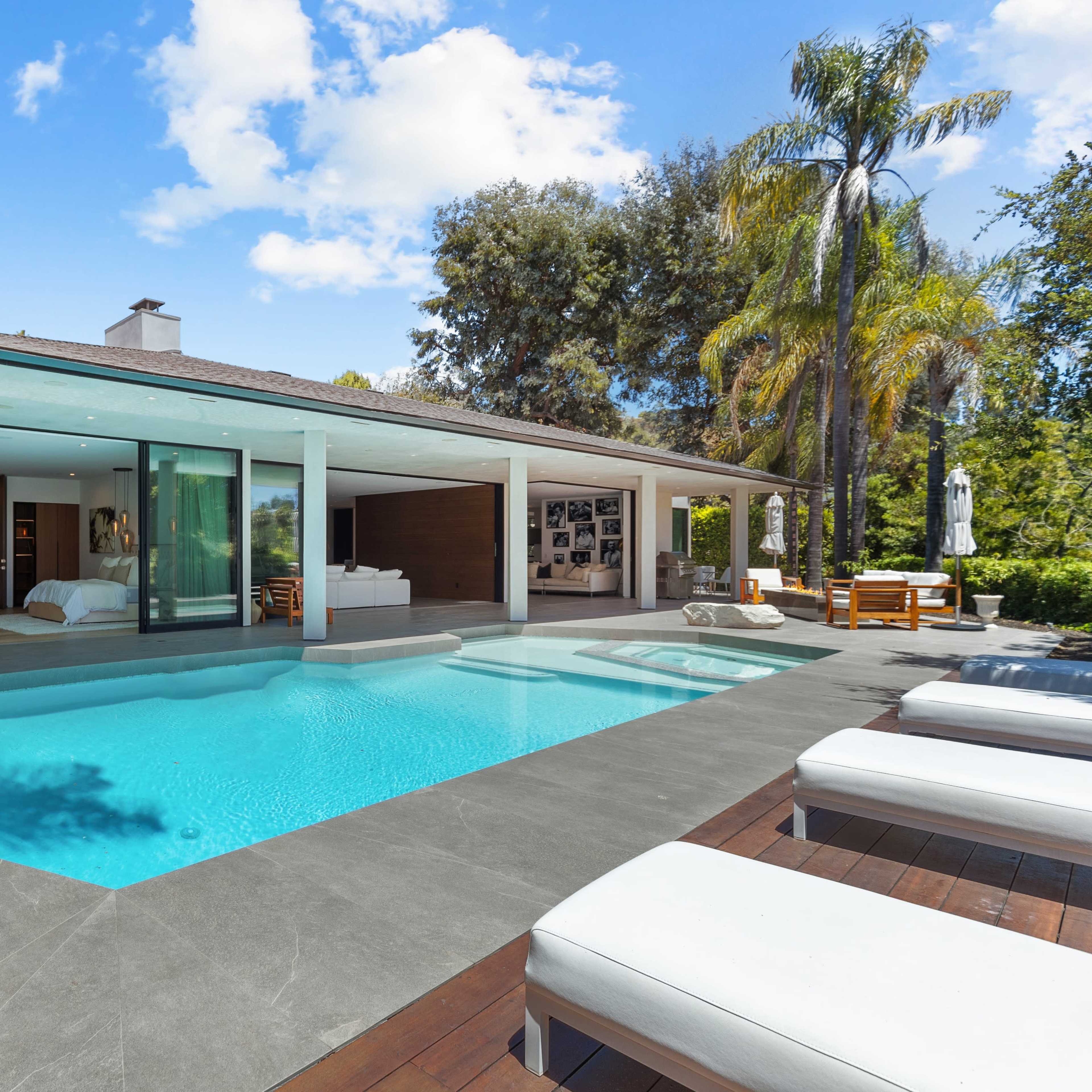 A modern one-story house features a landscaped yard with a pool and patio area, surrounded by palm trees.