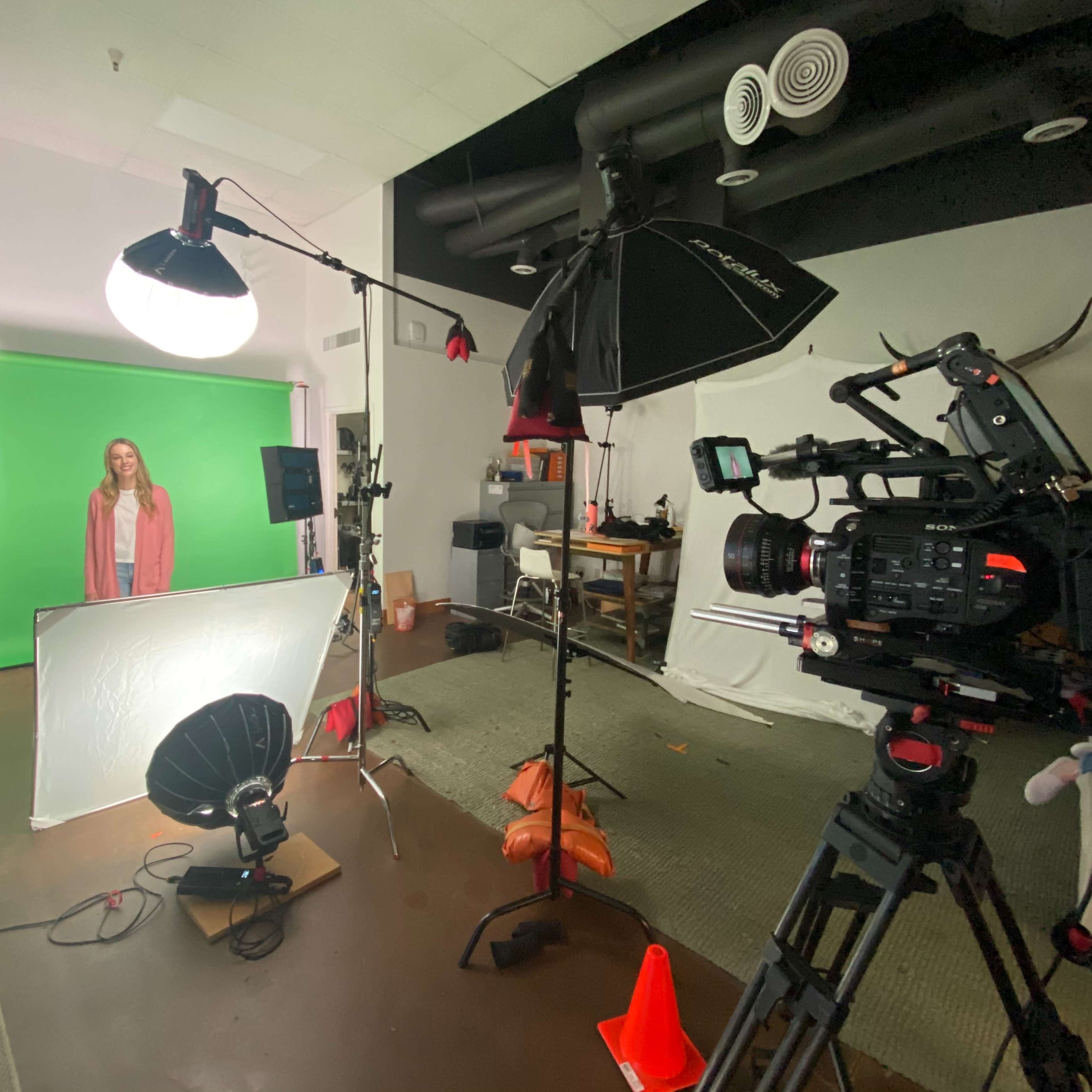 A woman stands in front of a green screen while various cameras and lighting equipment are set up around her.