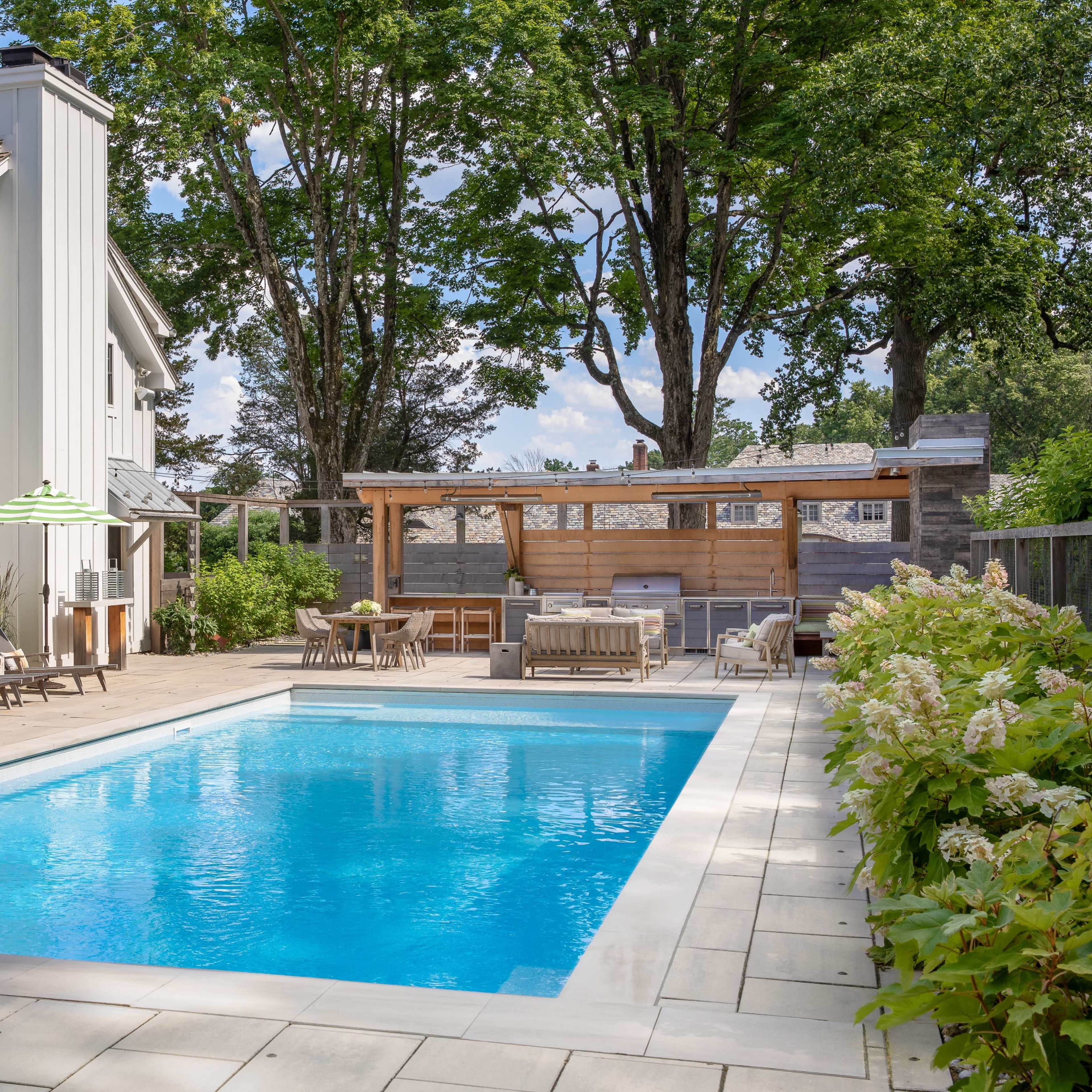 A backyard features a rectangular swimming pool surrounded by lounge chairs, a covered seating area, and lush greenery.